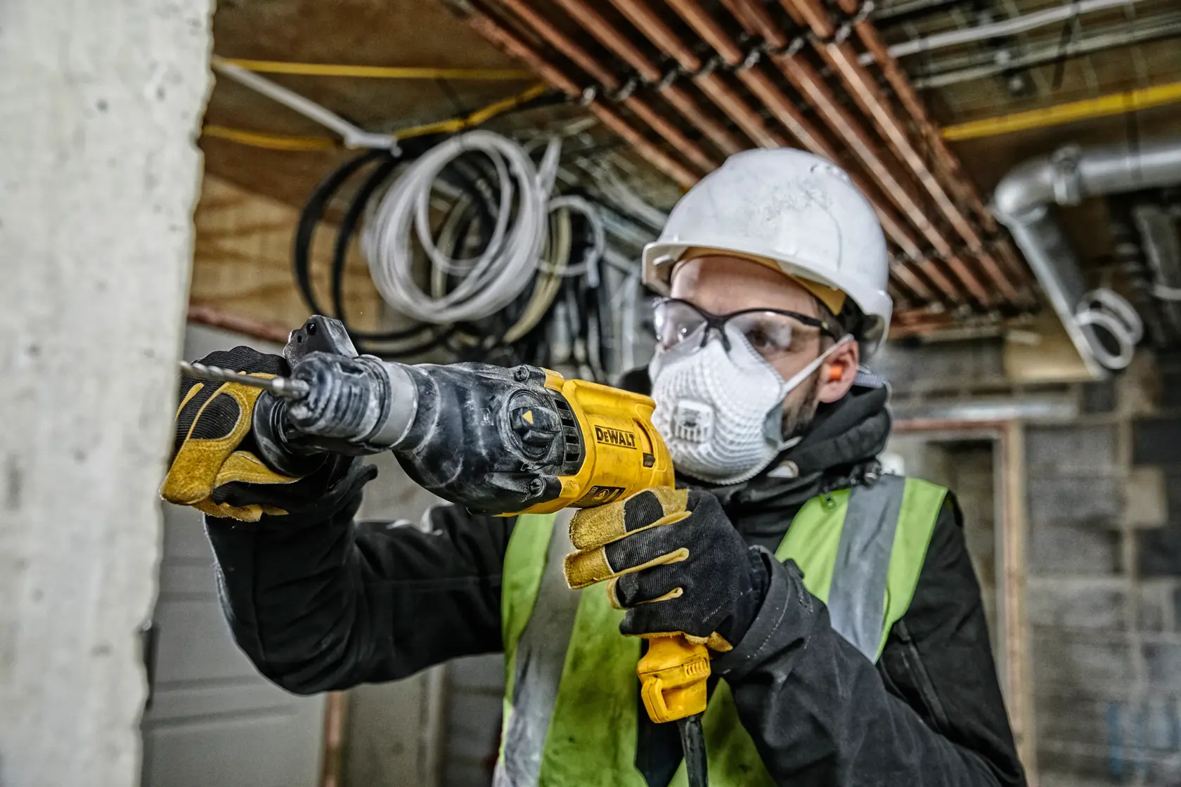 A construction worker wearing a hard hat, safety glasses, gloves, and a high-visibility vest is using a DEWALT D25033 rotary hammer drill at a building site. The background shows unfinished walls, pipes, and electrical wiring.