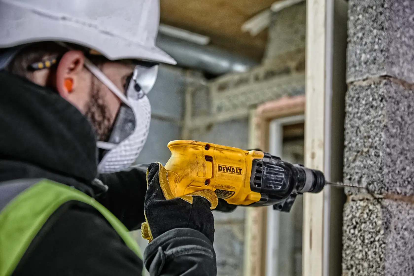 A person wearing safety equipment uses a yellow DEWALT power drill to drill into a concrete wall at a construction site.