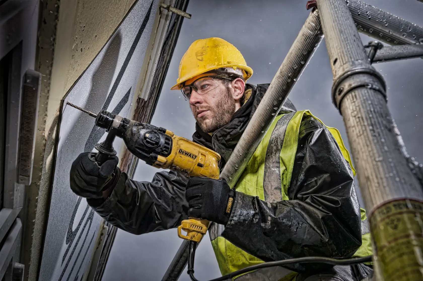 A construction worker wearing a yellow hard hat and safety vest is using a DEWALT hammer drill (model D25033) to drill into a wall while standing on scaffolding. The worker's face is blurred for privacy.