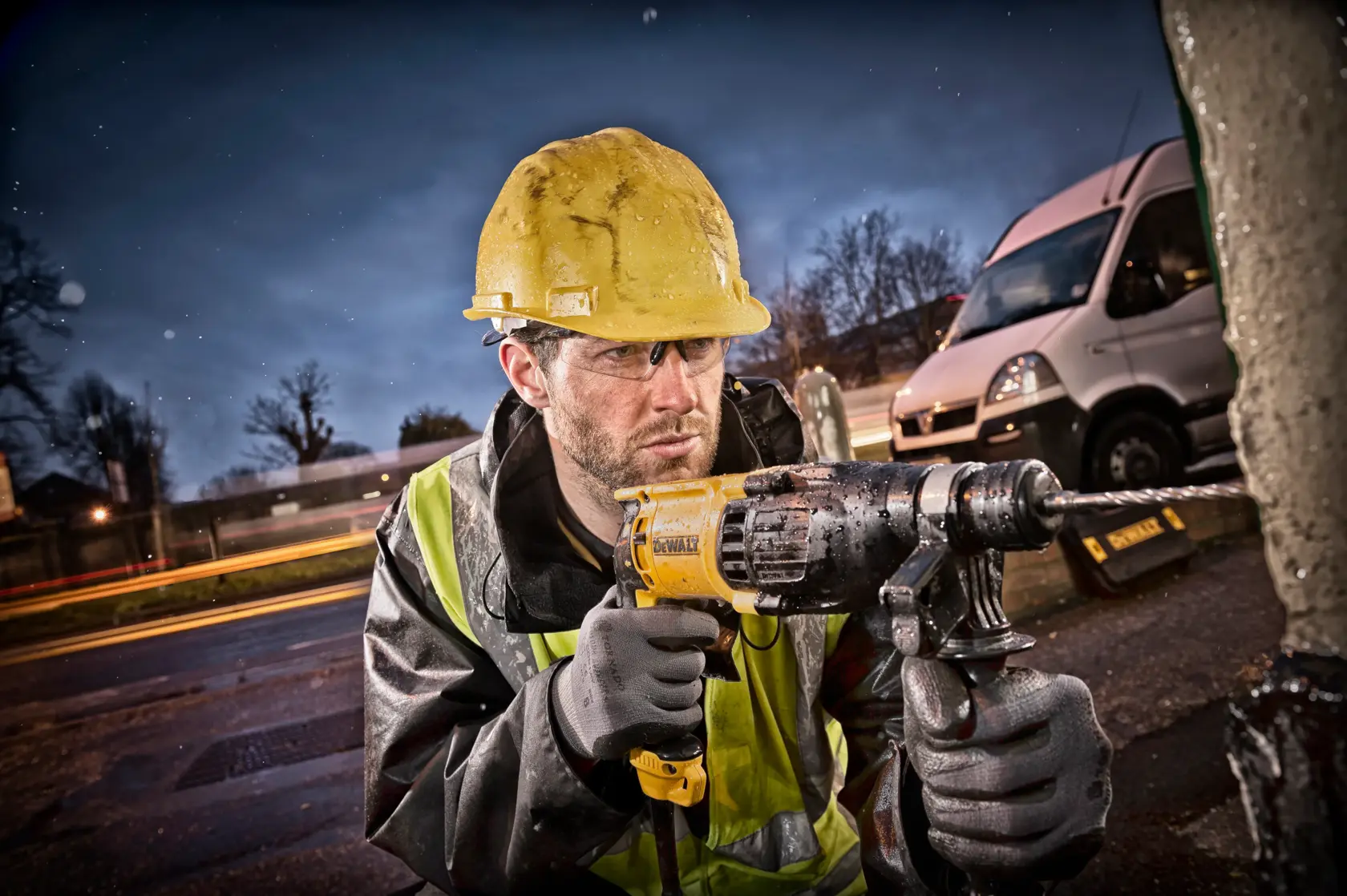 A construction worker wearing a yellow hard hat and reflective jacket uses a DEWALT hammer drill outdoors, with a white van and blurred lights in the background.