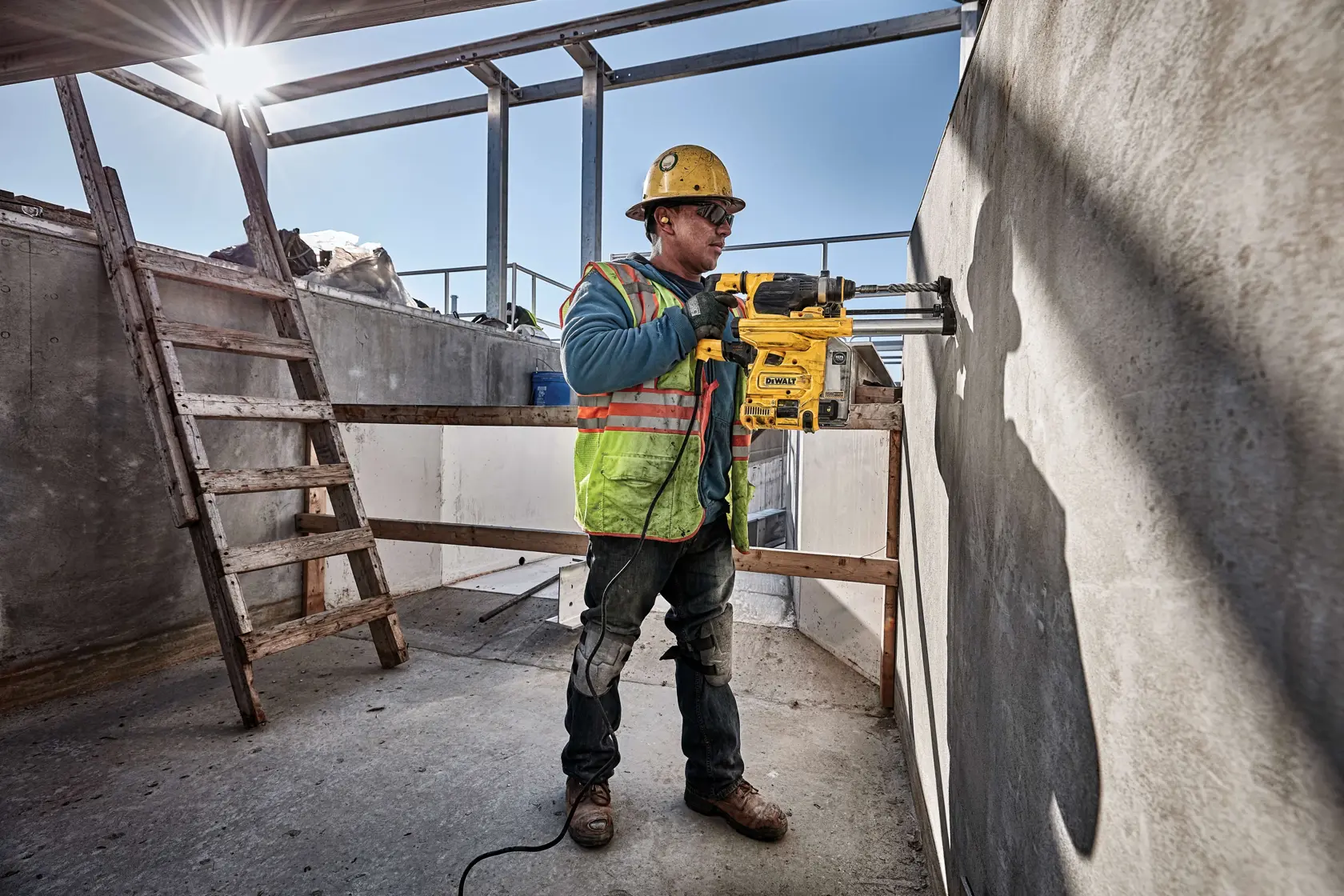 A construction worker wearing a safety helmet and high-visibility vest operates a yellow DEWALT rotary hammer drill, model D25333K, to bore a hole into a concrete wall at an outdoor construction site. The worker stands near a wooden ladder under a partly built structure with sunlight streaming in.