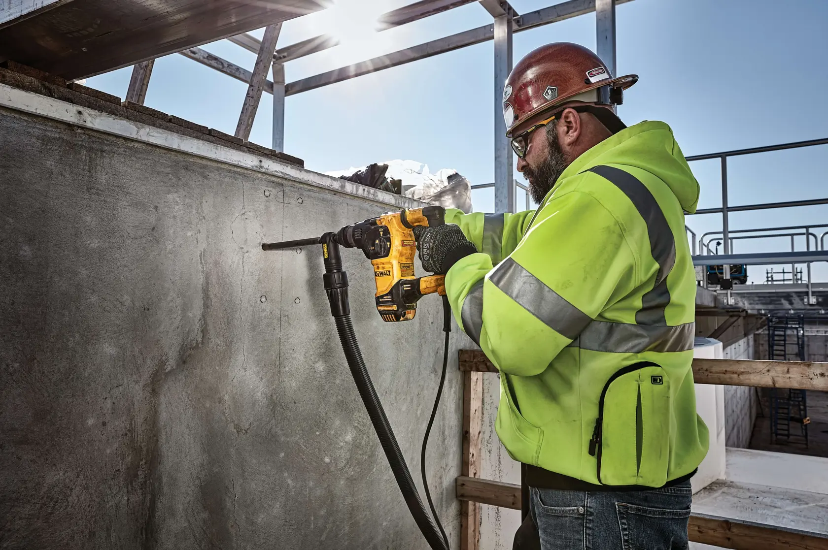 A person wearing a safety helmet and a high-visibility jacket is using a yellow DEWALT power tool to drill into a concrete wall at a construction site.