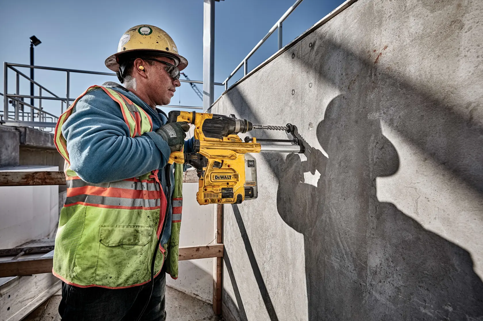 A construction worker wearing safety gear operates a DEWALT D25333K rotary hammer drill against a concrete wall at an outdoor job site.
