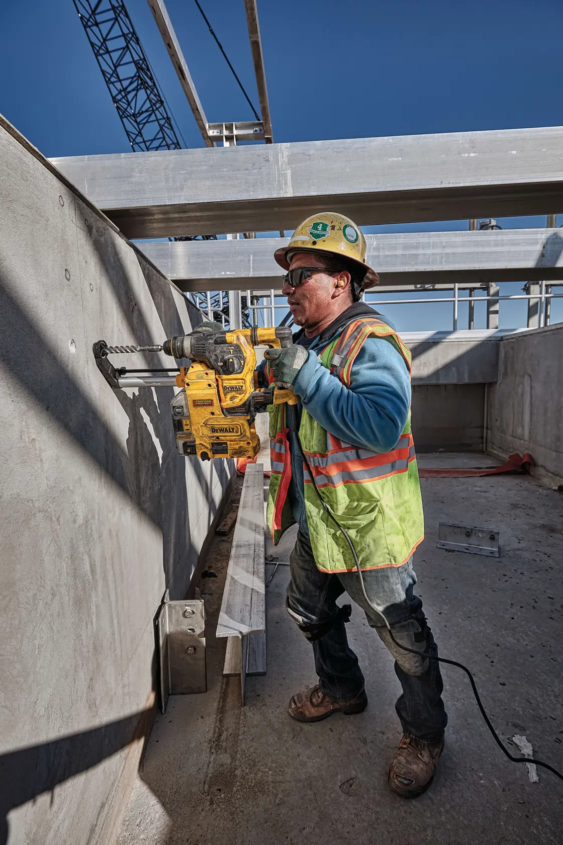 A construction worker wearing a safety vest and helmet is using a DEWALT D25333K rotary hammer drill to drill into a concrete wall at a construction site. The worker's face is blurred for privacy.