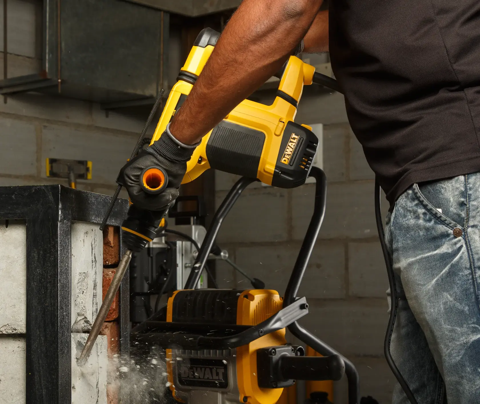 A person using a DEWALT rotary hammer drill to chisel concrete in an industrial setting. Dust and debris are visible around the tool during operation.