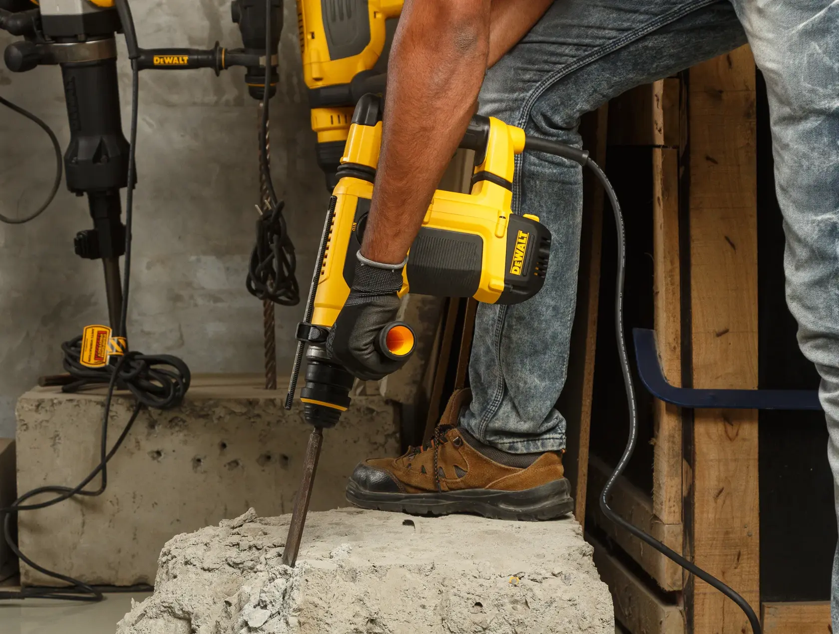 A person using a DEWALT rotary hammer drill (model D25413K) to break concrete in a construction setting. The person is wearing gloves, jeans, and brown work boots. The tool is plugged in and other power tools are visible in the background.