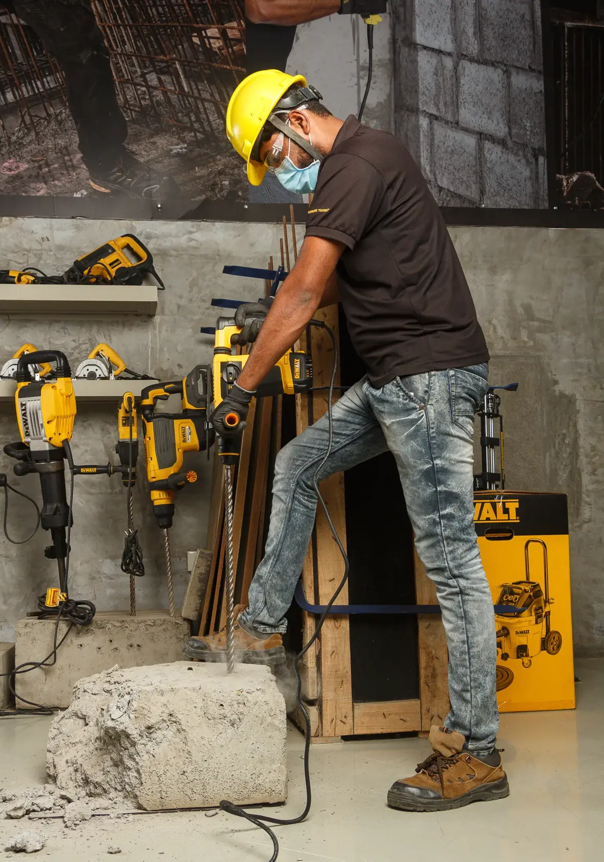A person wearing safety gear uses a DEWALT hammer drill to break a large concrete block in a workshop. Other DEWALT power tools are visible in the background, along with construction materials.