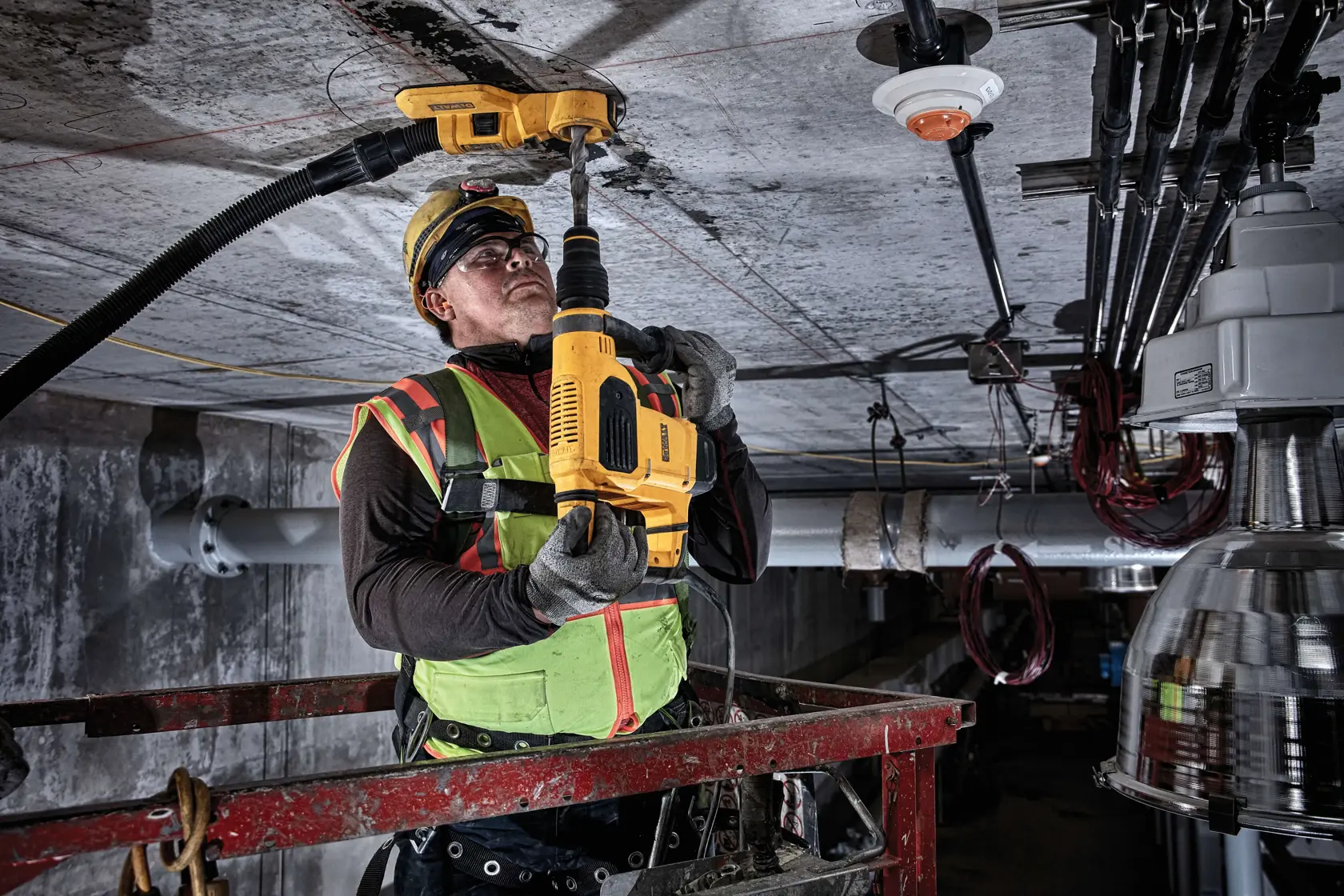 Worker uses a DEWALT drill on a ceiling at a construction site, wearing safety gear and a high-visibility vest.