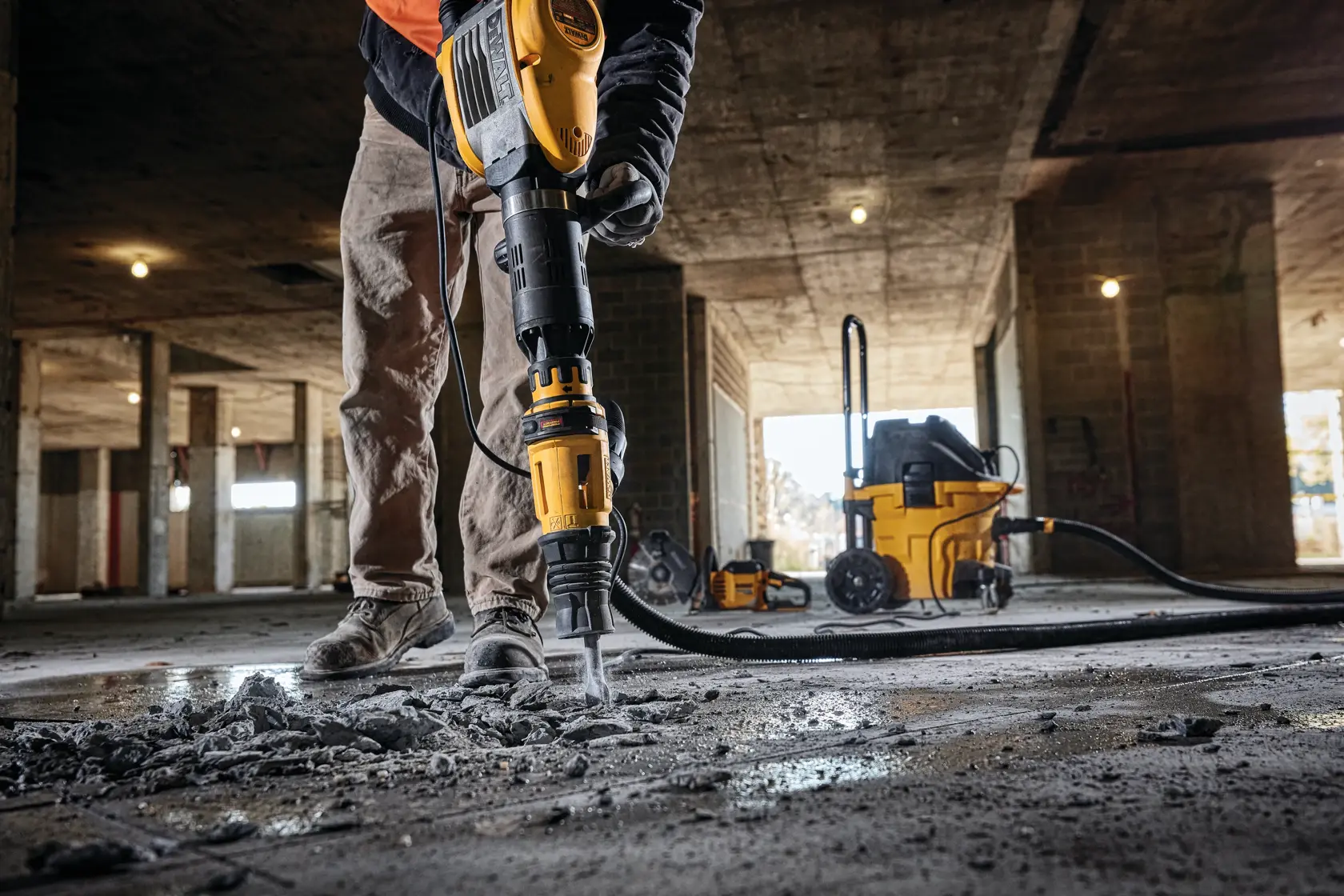 A person using a DEWALT D25951K demolition hammer to break concrete on a construction site, with debris scattered on the floor and additional DEWALT equipment visible in the background.