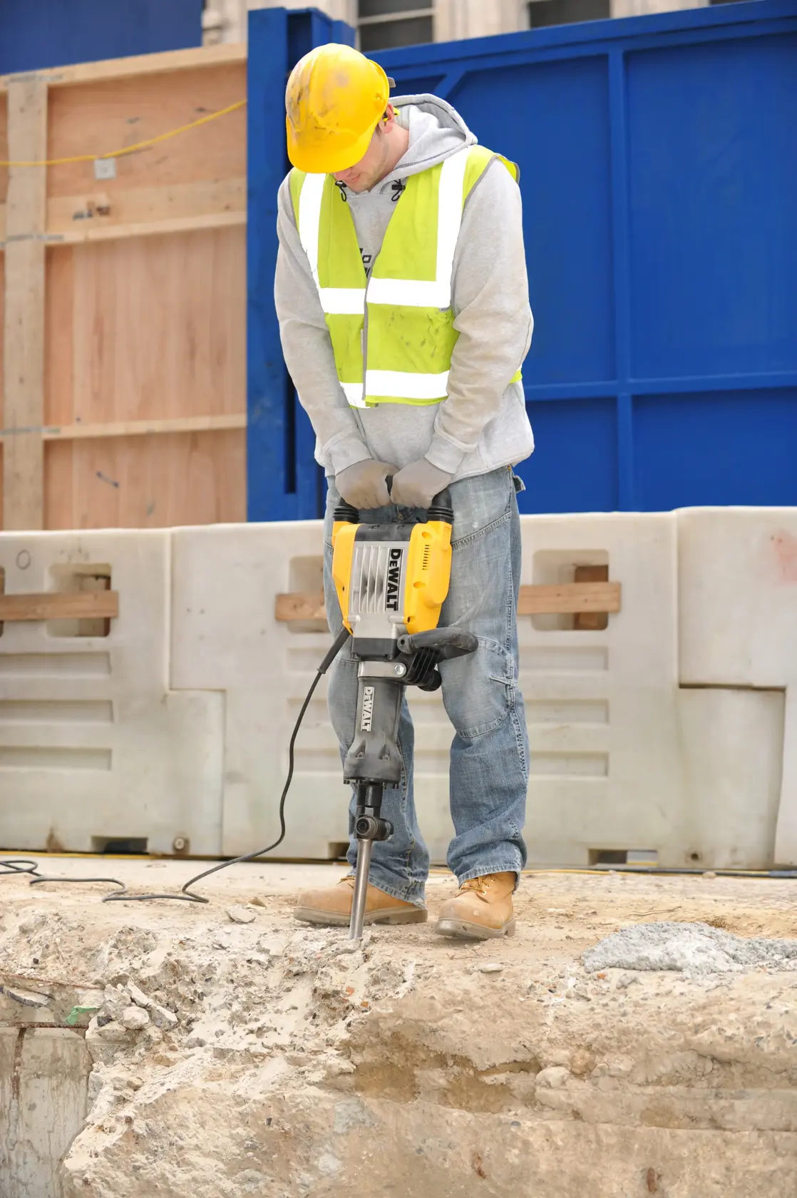 A person wearing a yellow hard hat, safety gloves, and a reflective safety vest is using a DEWALT electric jackhammer (model D25961K) to break concrete at an outdoor construction site.