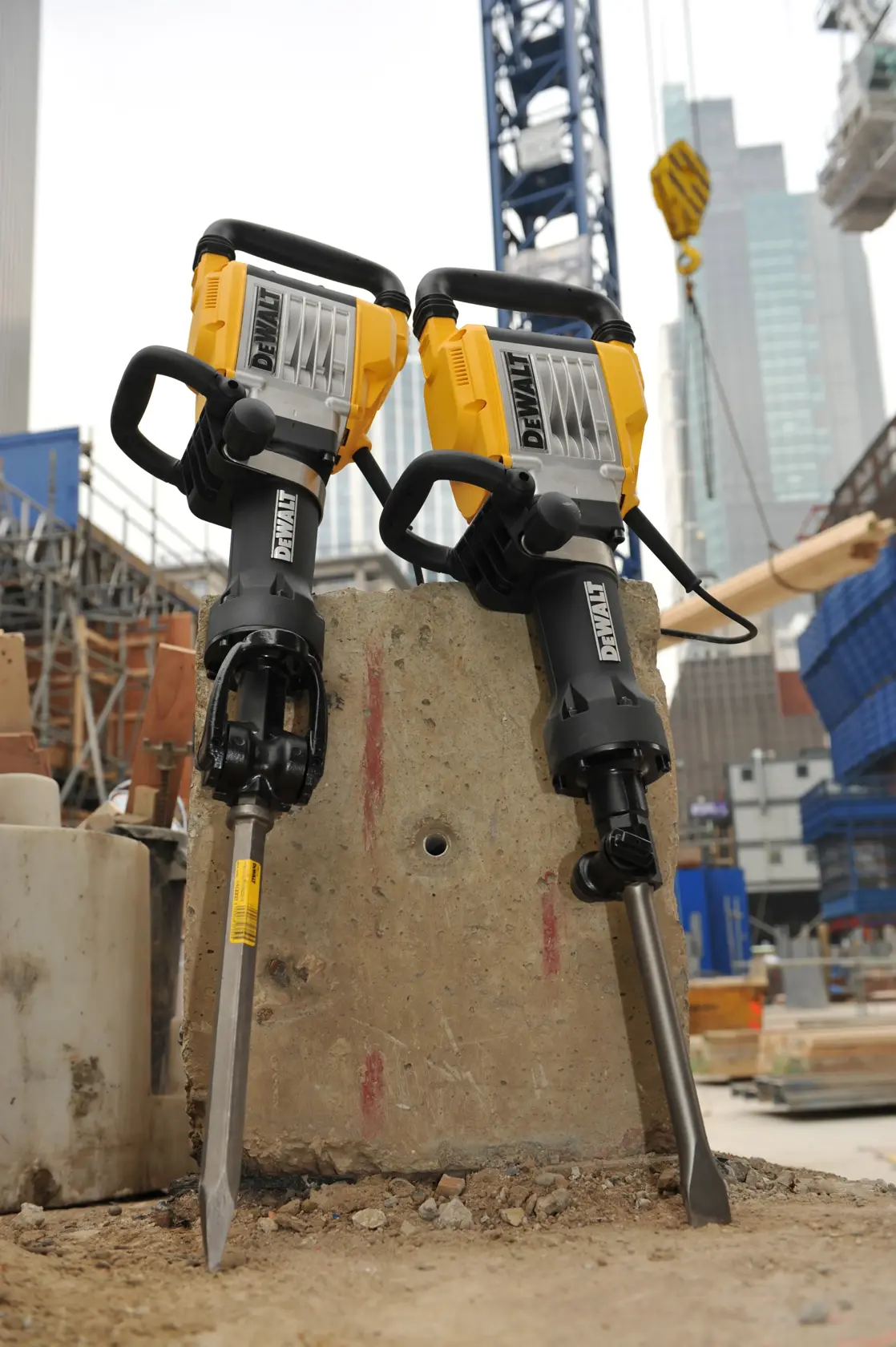 Two DEWALT electric demolition hammers are positioned upright on a concrete block at a construction site, with buildings and cranes visible in the background.