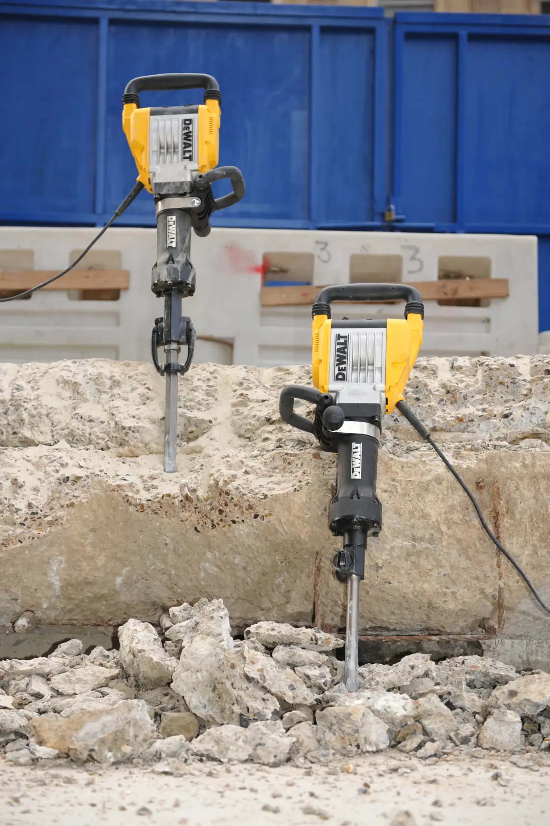 Two DEWALT electric demolition hammers are shown breaking up a large concrete slab, with rubble and broken pieces of concrete on the ground. The tools are positioned vertically and connected by cables, in an outdoor construction setting.