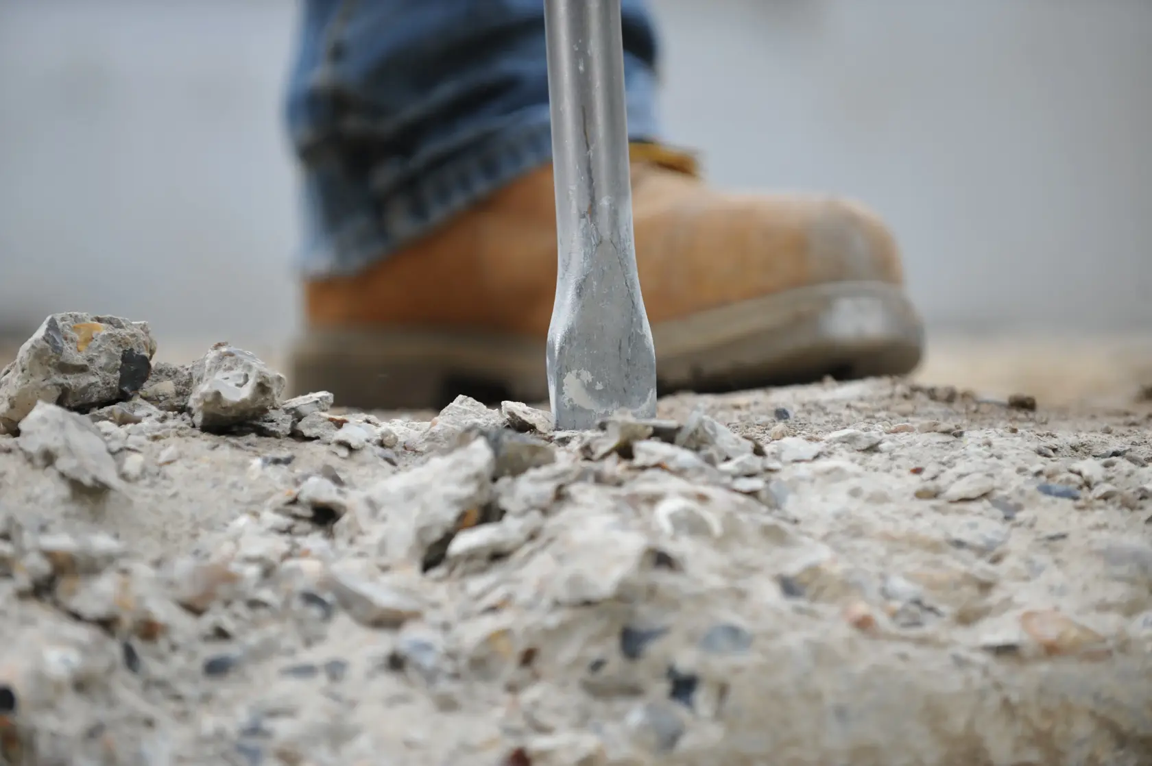Close-up view of a construction worker's boot and a chisel breaking concrete on a worksite.