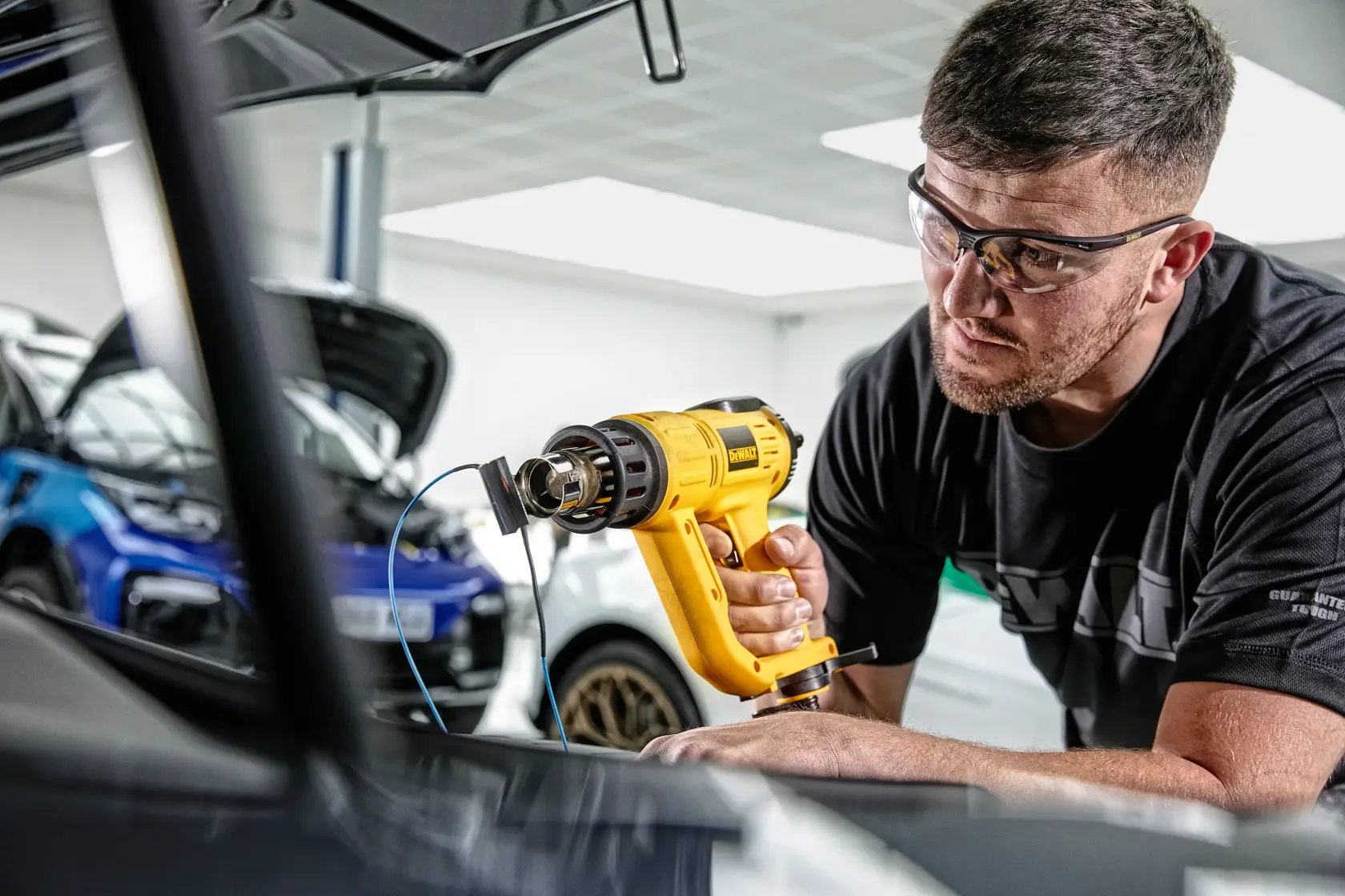A person uses a DEWALT D26414 yellow heat gun in a garage or workshop, with cars visible in the background. The person’s face is blurred for privacy.