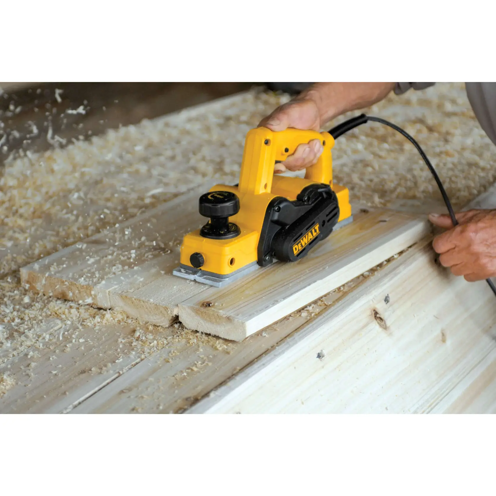 A person using a yellow DEWALT electric hand planer to smooth wooden boards, with wood shavings scattered around the workspace.