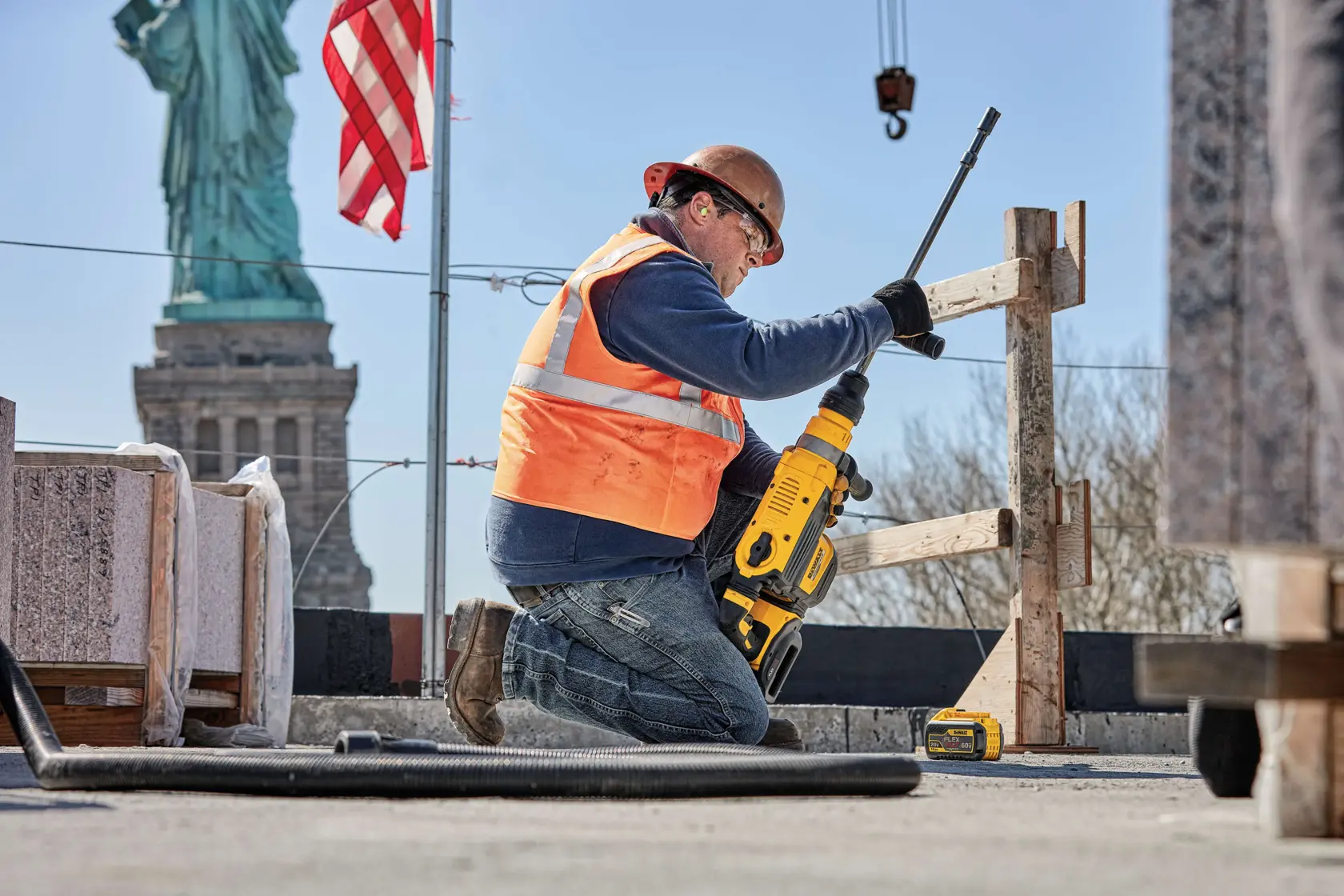 A construction worker wearing an orange safety vest and helmet uses a yellow DEWALT power tool for drilling at a construction site. The worker is kneeling near wooden structures, with the DEWALT DCB612 battery visible beside them. The Statue of Liberty and an American flag are visible in the background.