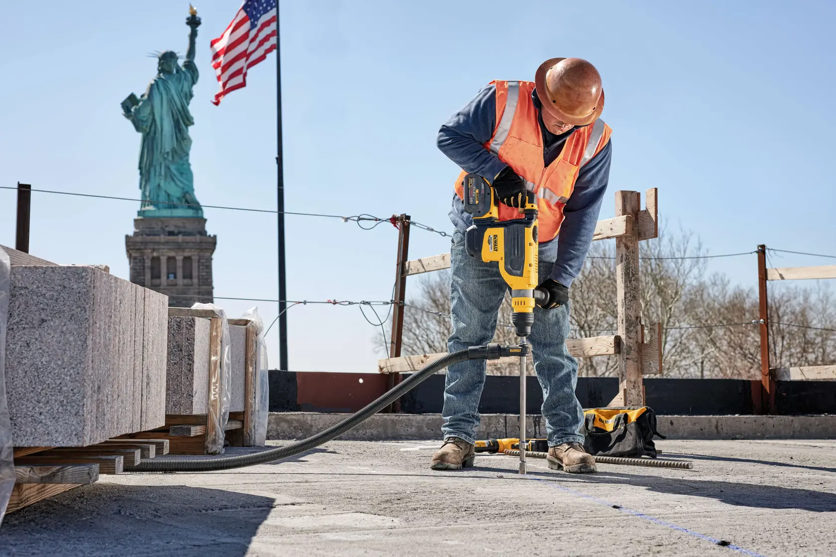 A construction worker wearing a hard hat and safety vest uses a DEWALT power tool to drill into concrete at a construction site. The Statue of Liberty and an American flag are visible in the background.