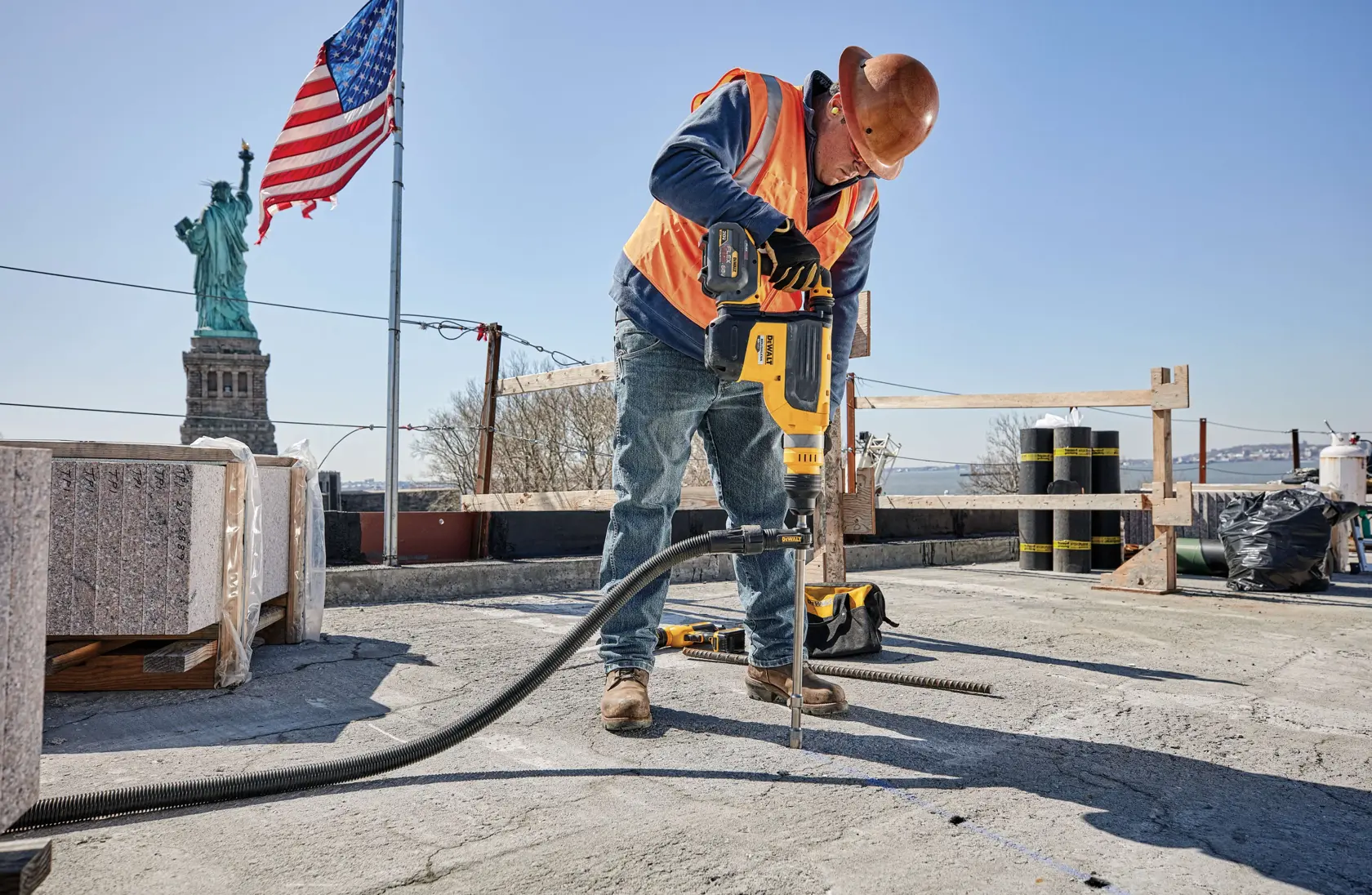 Worker wearing a hard hat and orange safety vest using a yellow DEWALT power tool to drill concrete at a construction site with the Statue of Liberty and an American flag in the background.