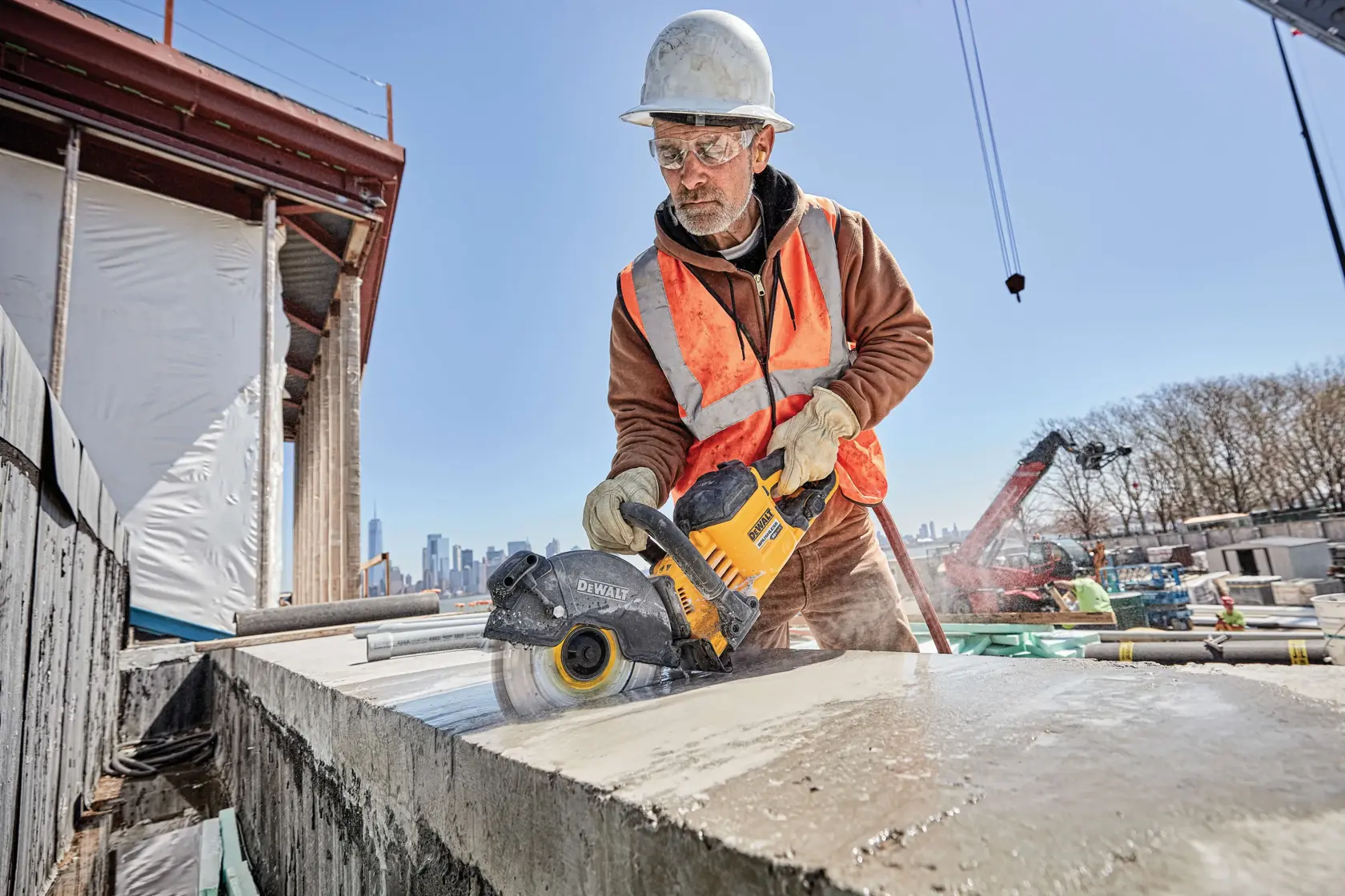 A construction worker wearing a safety helmet and orange reflective vest is using a DEWALT power tool to cut through a concrete slab at a construction site. The worker's face is blurred, and there is visible dust from the cutting process. The background shows structural beams, equipment, and other workers.