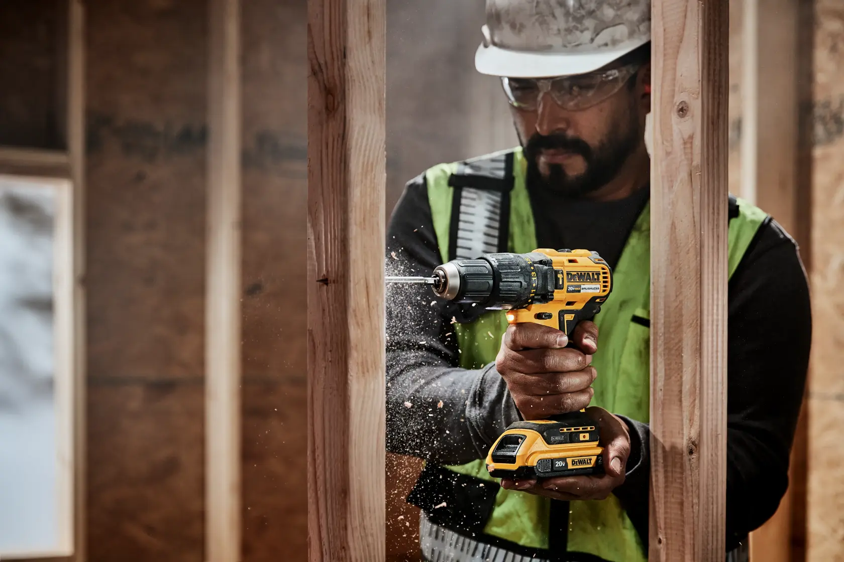 A person wearing a safety vest and helmet is using a DEWALT cordless power drill to make a hole in a wooden beam at a construction site. Wood shavings are visible around the drill bit.