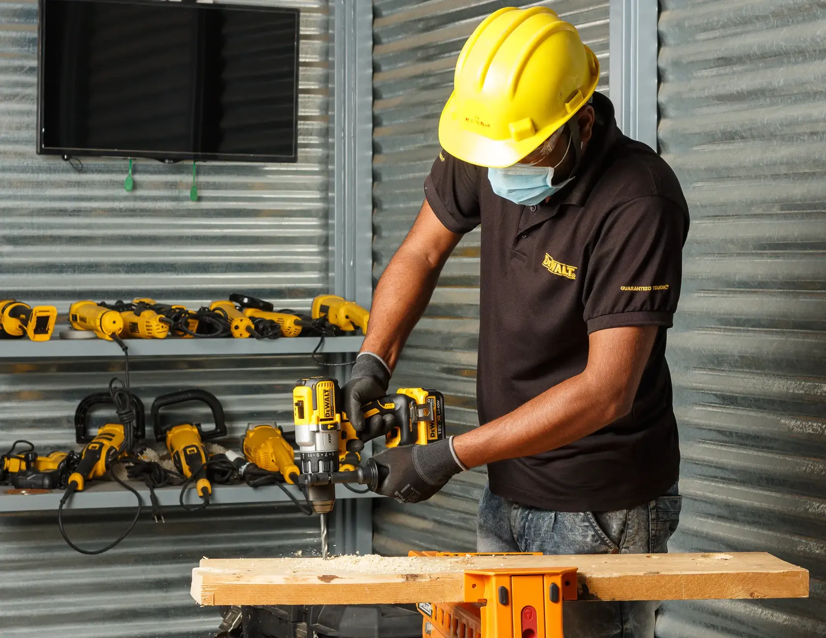 Worker using DEWALT DCD996NT drill on wood, wearing safety gear in workshop with other DEWALT tools in background.