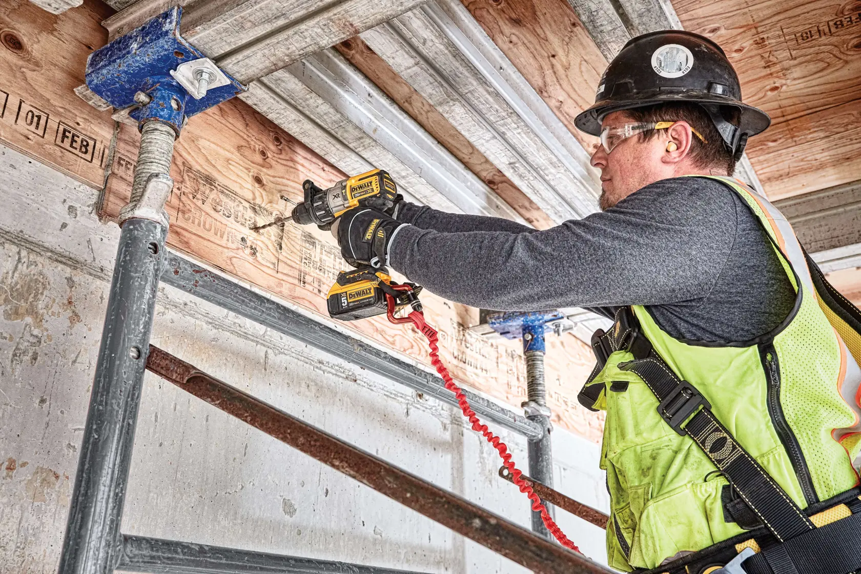 A construction worker wearing a safety vest and hard hat uses a DEWALT cordless drill (SKU: DCD996P2) to drill into wooden beams on a construction site, standing on scaffolding.