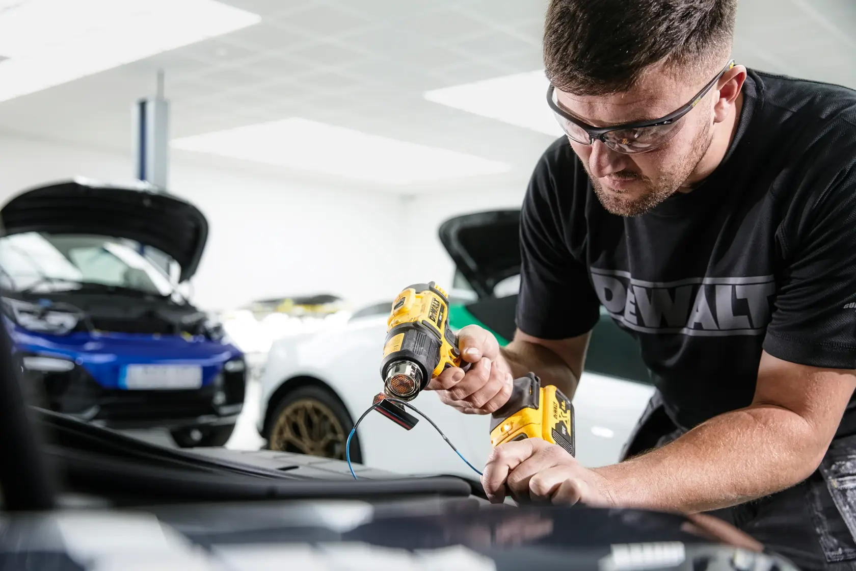 A person using a DEWALT cordless heat gun (model DCE530N) on a car part, with the face blurred for privacy. The individual is wearing a DEWALT-branded shirt and is working in a garage with cars in the background.