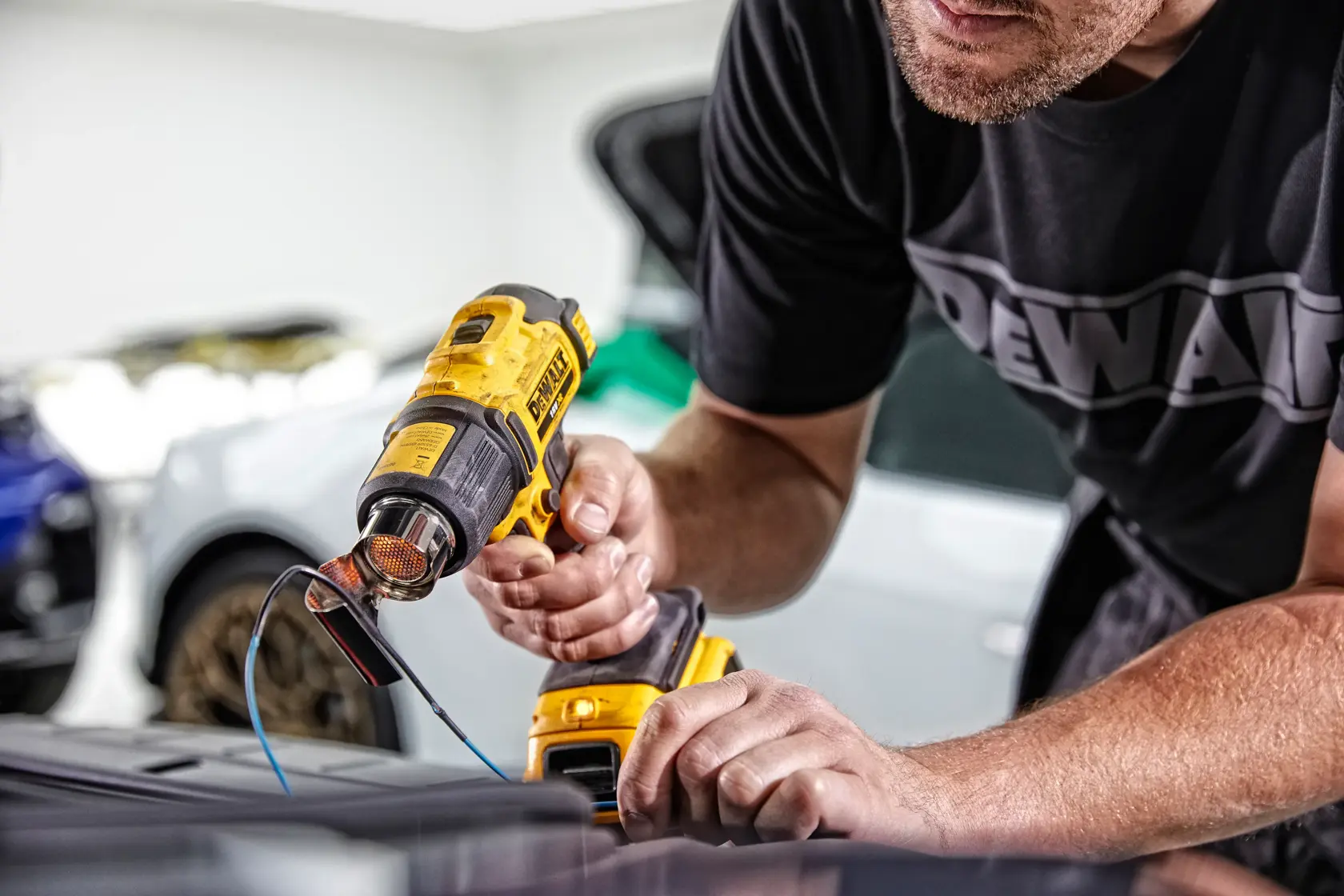 A person uses a DEWALT DCE530N cordless heat gun to work on a surface, with wires and cars visible in the background.