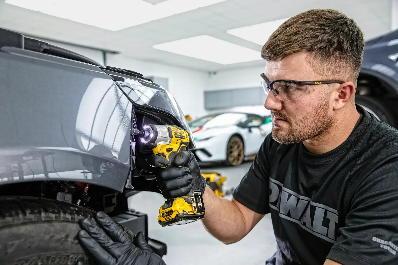 A person wearing black gloves uses a yellow DEWALT cordless drill to work on the front end of a gray car in a garage. The face of the person is blurred for privacy and there are other cars in the background.