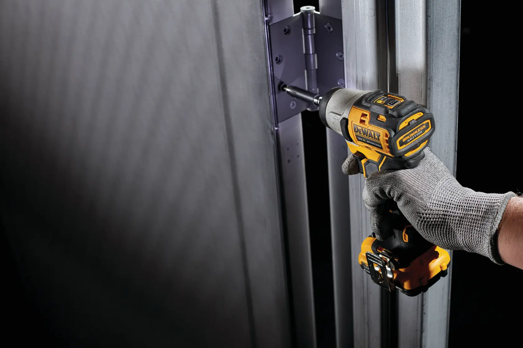 A person wearing a grey work glove uses a DEWALT cordless impact driver to install or tighten screws on a metal door hinge.