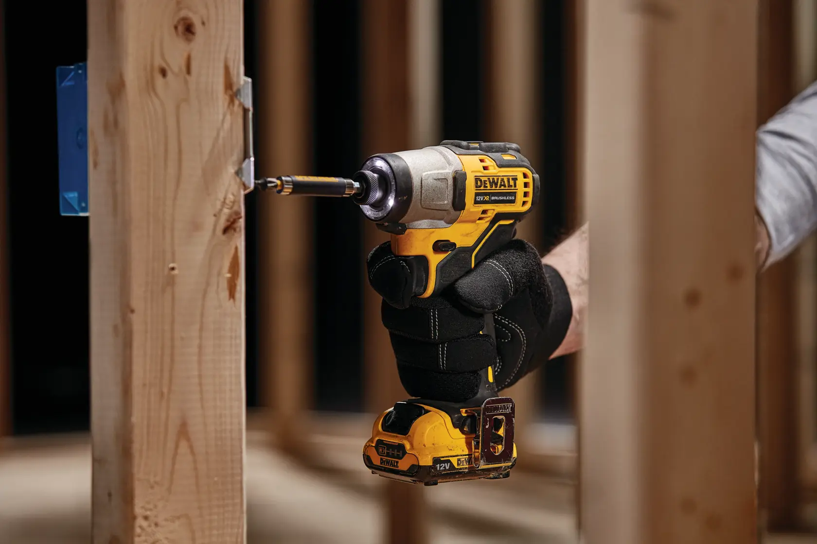 A person wearing black gloves is using a DEWALT cordless impact driver to drive a screw into a wooden frame in a construction setting.