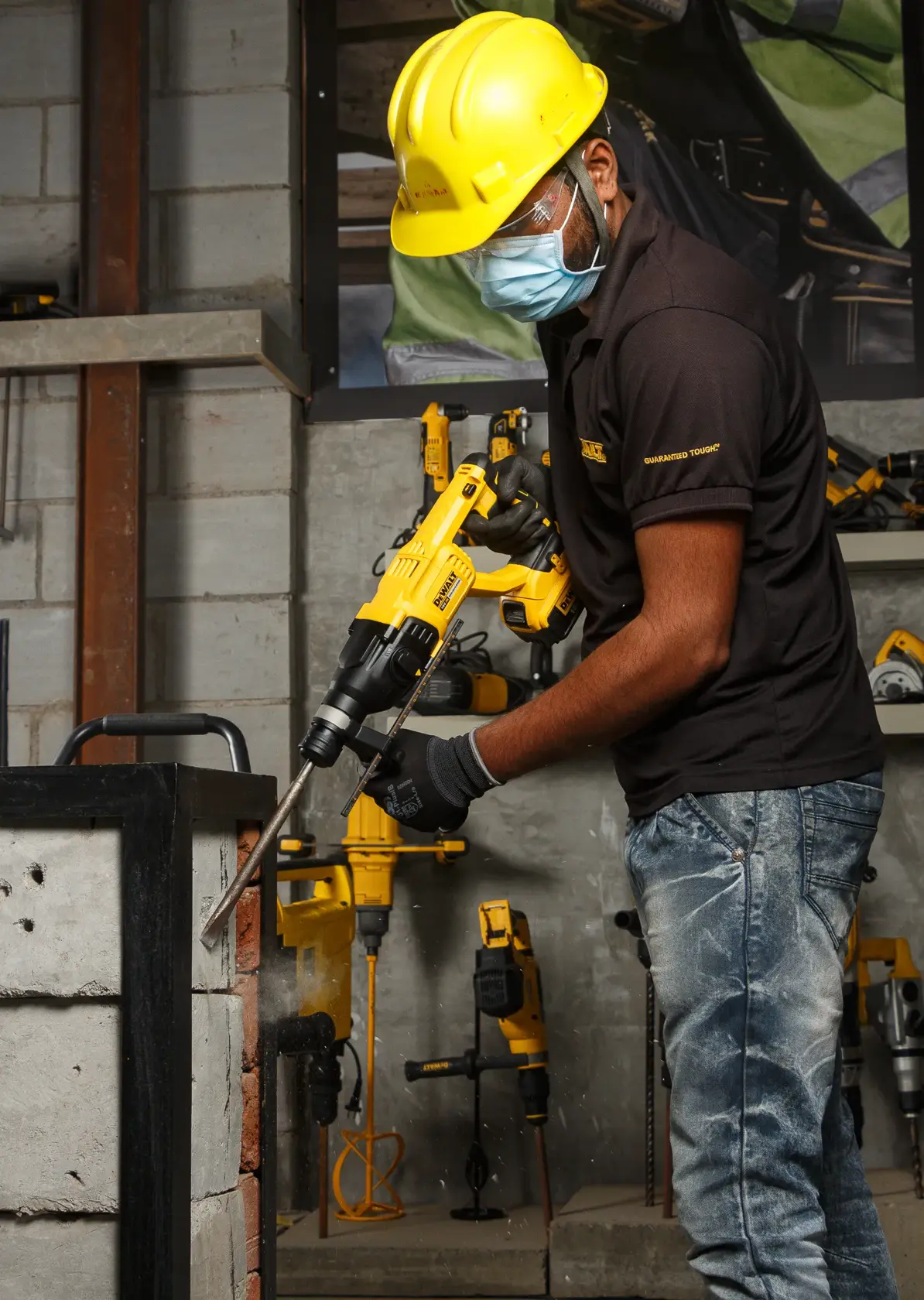 Worker uses DEWALT DCH133NT rotary hammer drill on a concrete wall. Protective gear and tools are visible.