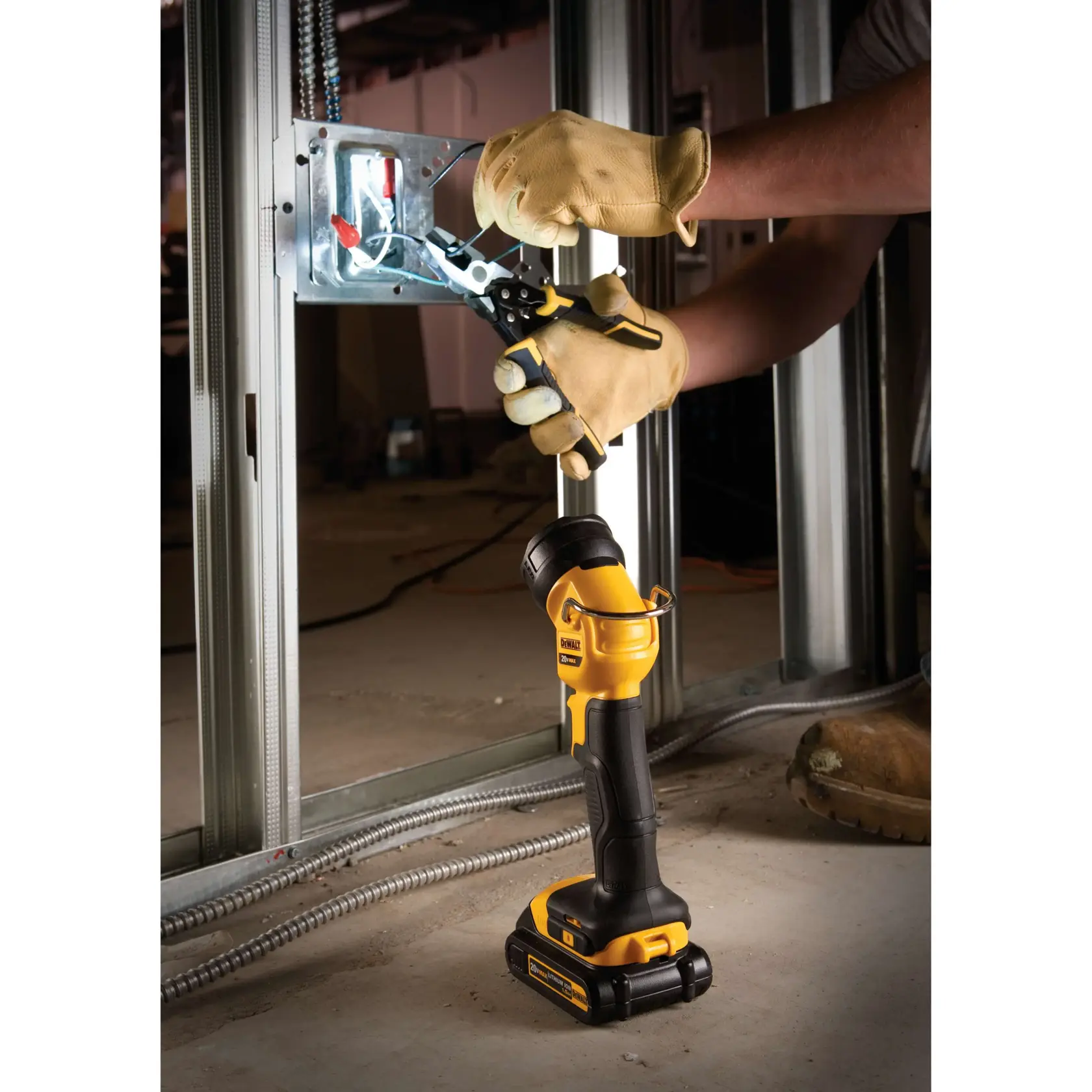 A DEWALT DCL040 portable LED work light is placed on the floor, illuminating an electrical box while a person wearing work gloves uses pliers to work on wiring inside a metal wall frame.