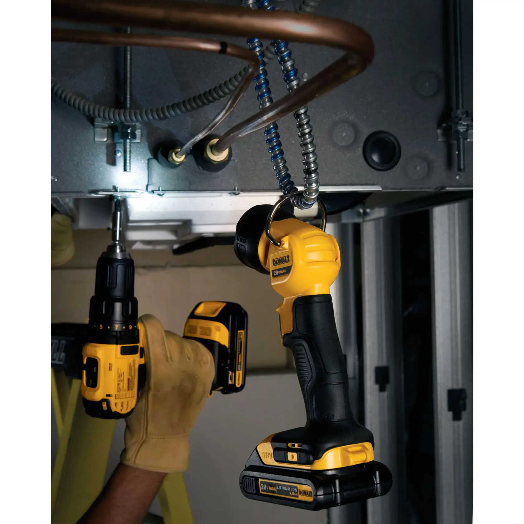 A DEWALT cordless LED work light (model DCL040) is hanging from a metal fixture, illuminating a workspace. Nearby, a person wearing tan work gloves uses a DEWALT cordless power drill to work on metal equipment and piping.