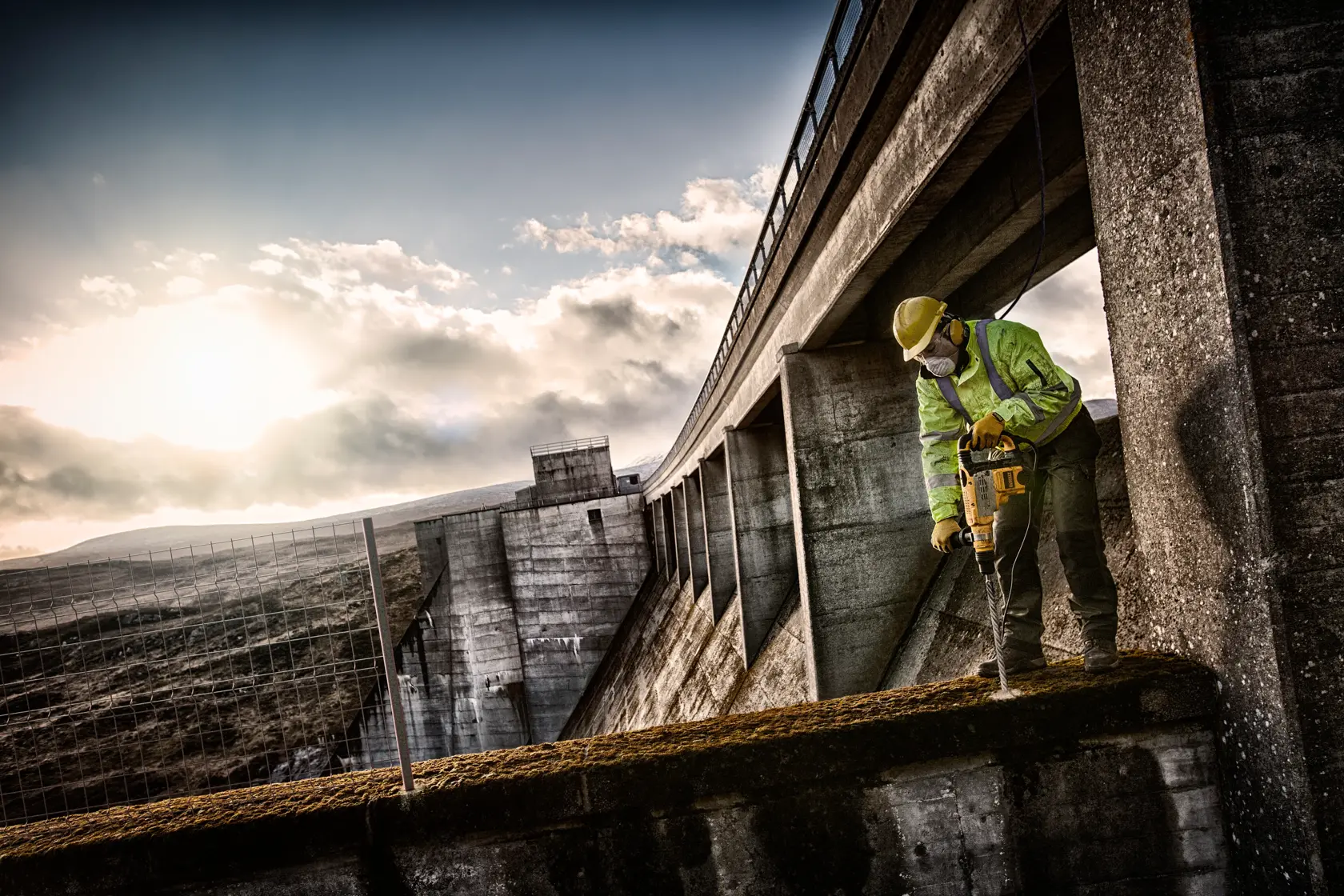 Worker using DEWALT drill on concrete structure outdoors with safety gear at sunset.