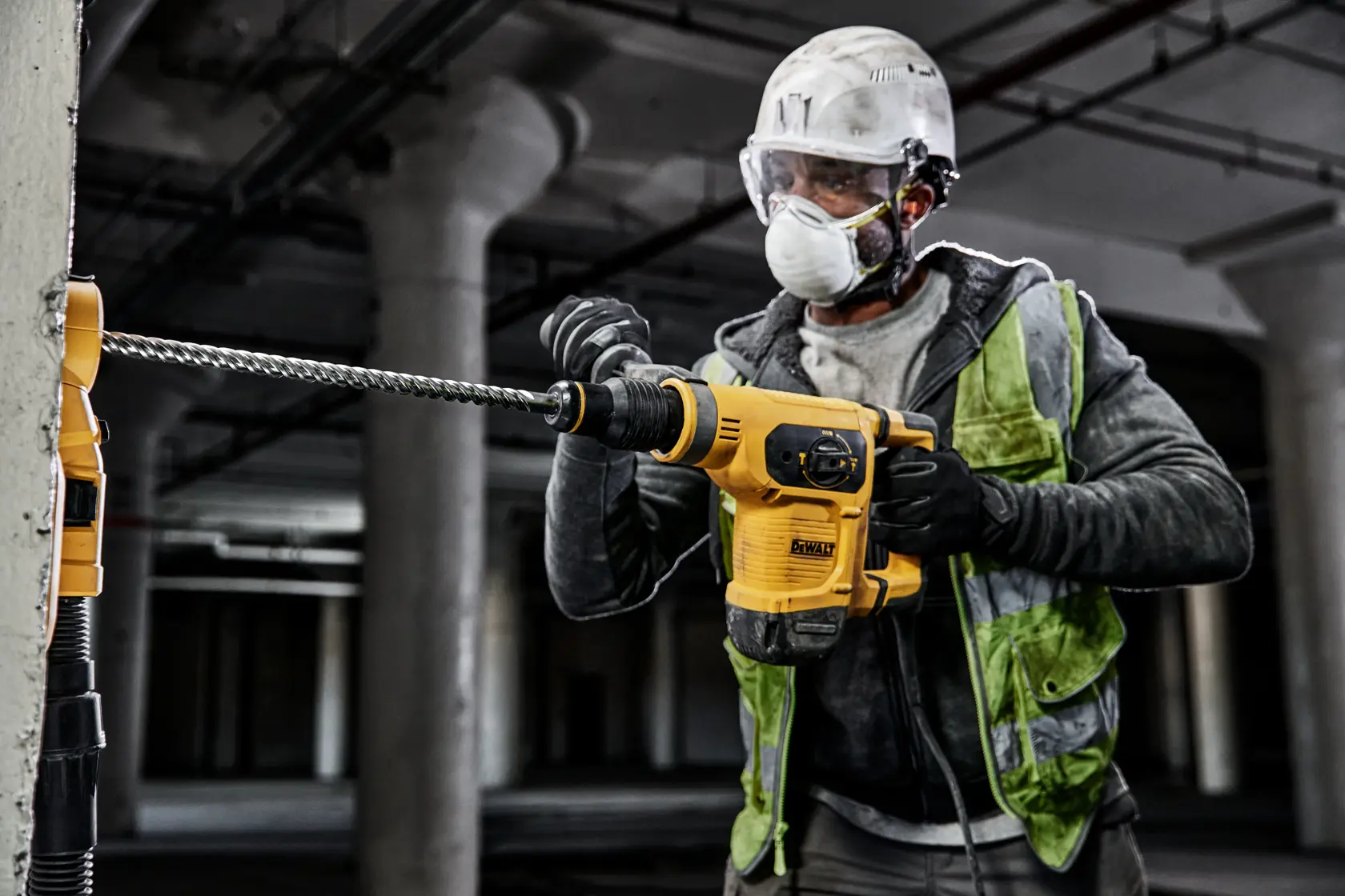 Worker drills concrete wall with DEWALT rotary hammer. Wearing safety gear and reflective vest at construction site.