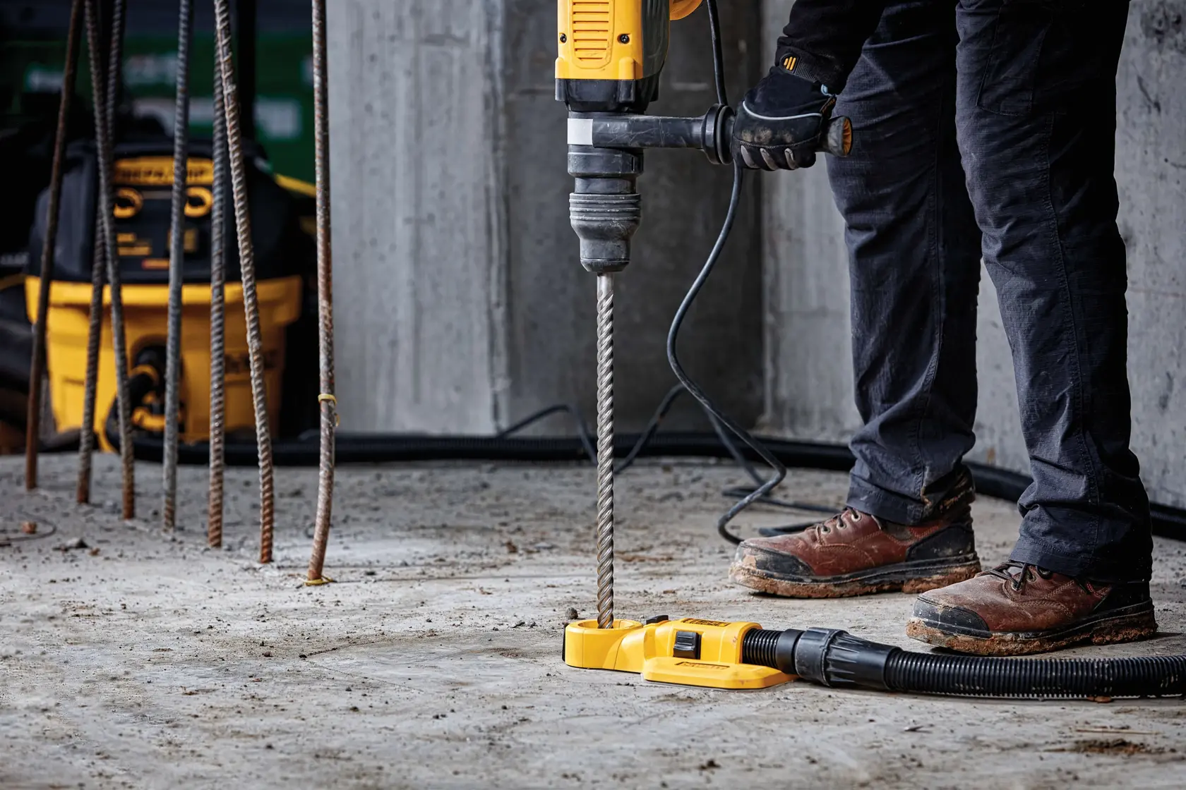 Person using DEWALT hammer drill with dust extractor on a concrete floor at a construction site.