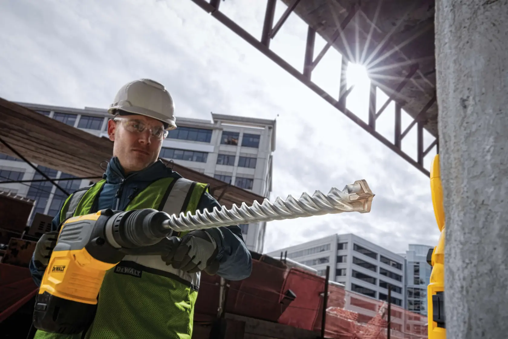 Worker using a DEWALT drill with a large bit at a construction site, wearing safety gear, with buildings in background.