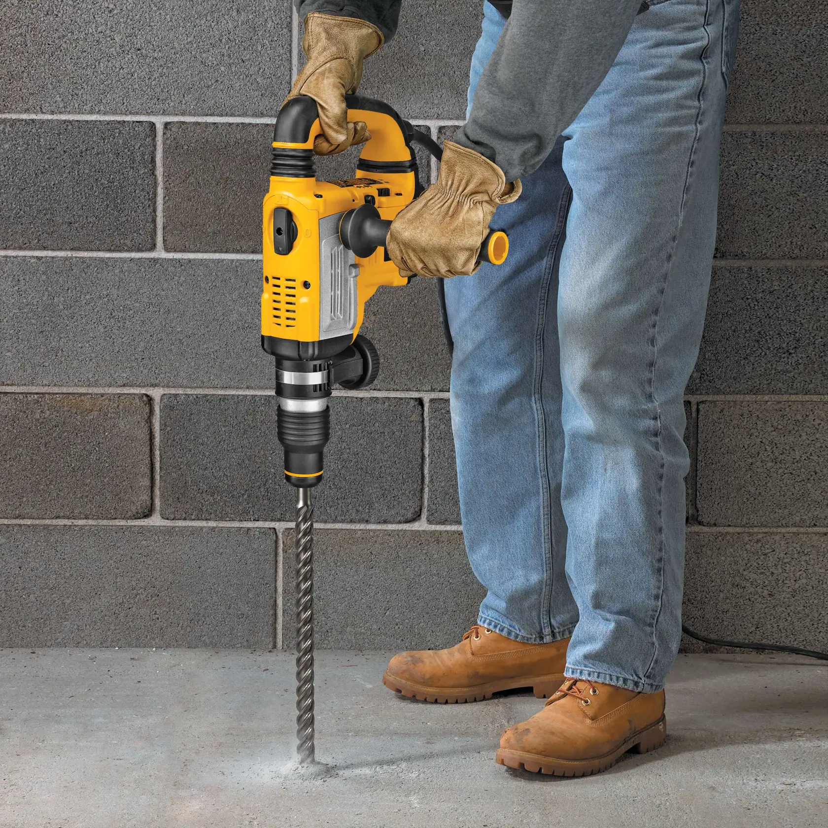 Person drills concrete floor with a yellow DEWALT rotary hammer drill, wearing gloves and boots.