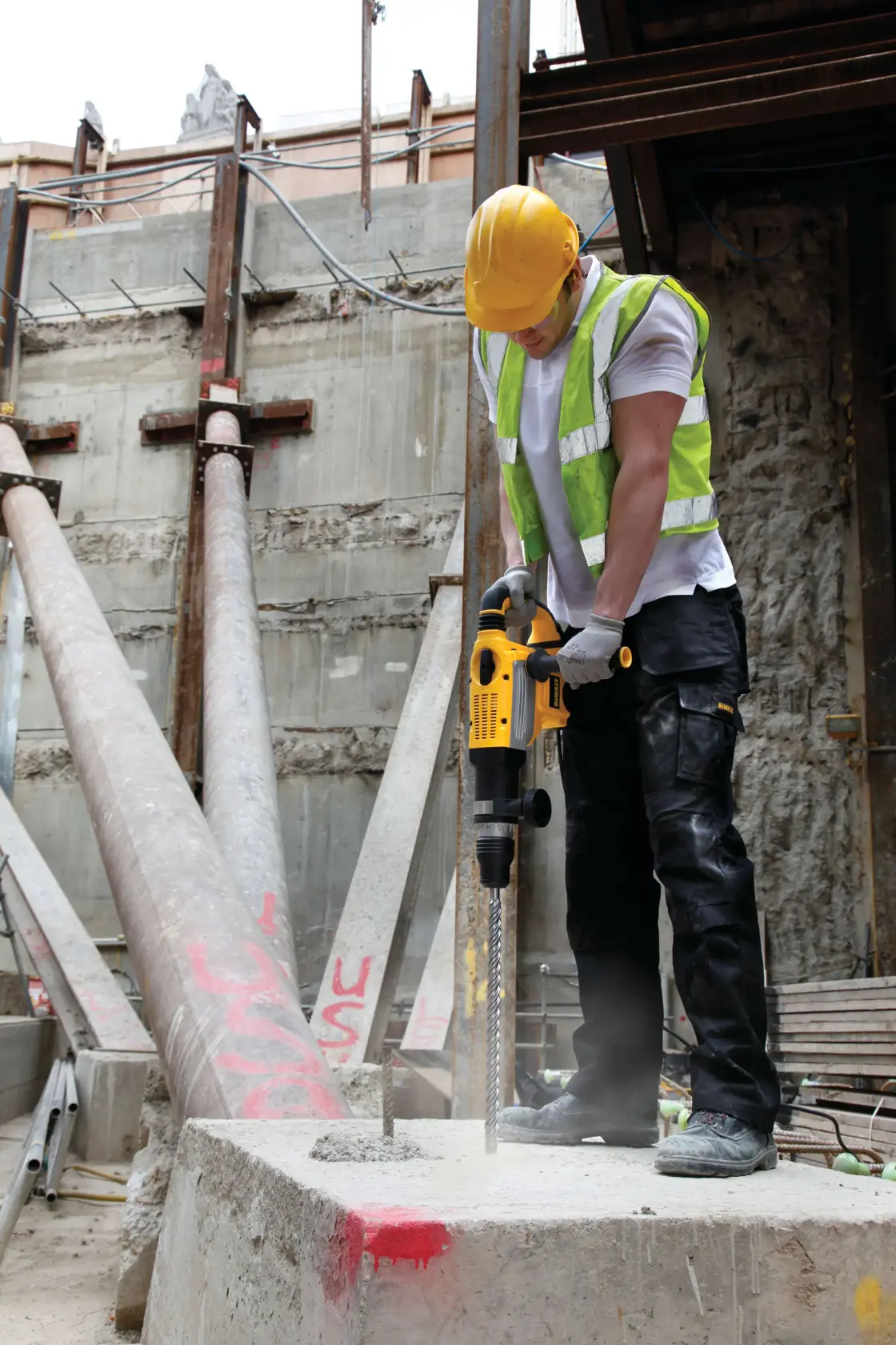 Worker uses yellow DEWALT drill on concrete at construction site. Wearing safety gear. DT9450 SKU visible.