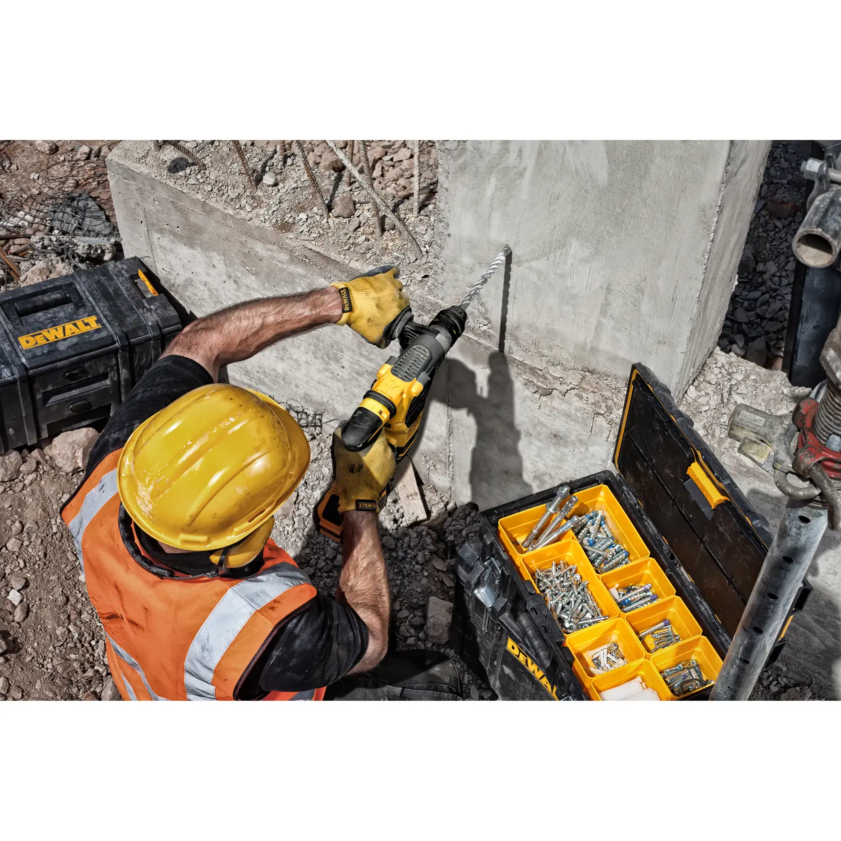 Worker drills concrete with a DEWALT tool, using screws from an organized yellow-black box on a construction site.