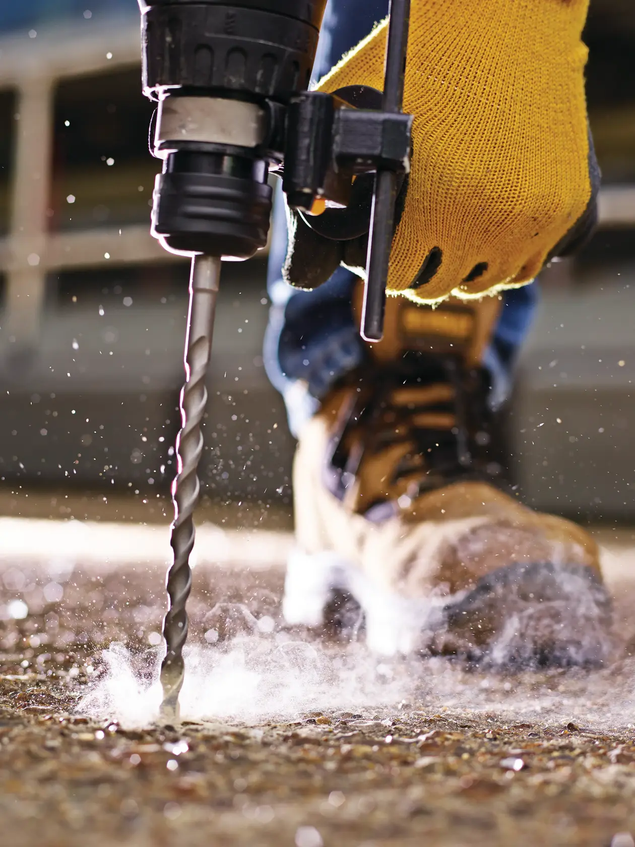 Close-up of a gloved hand using a drill bit on concrete, dust rising, work boot in background.