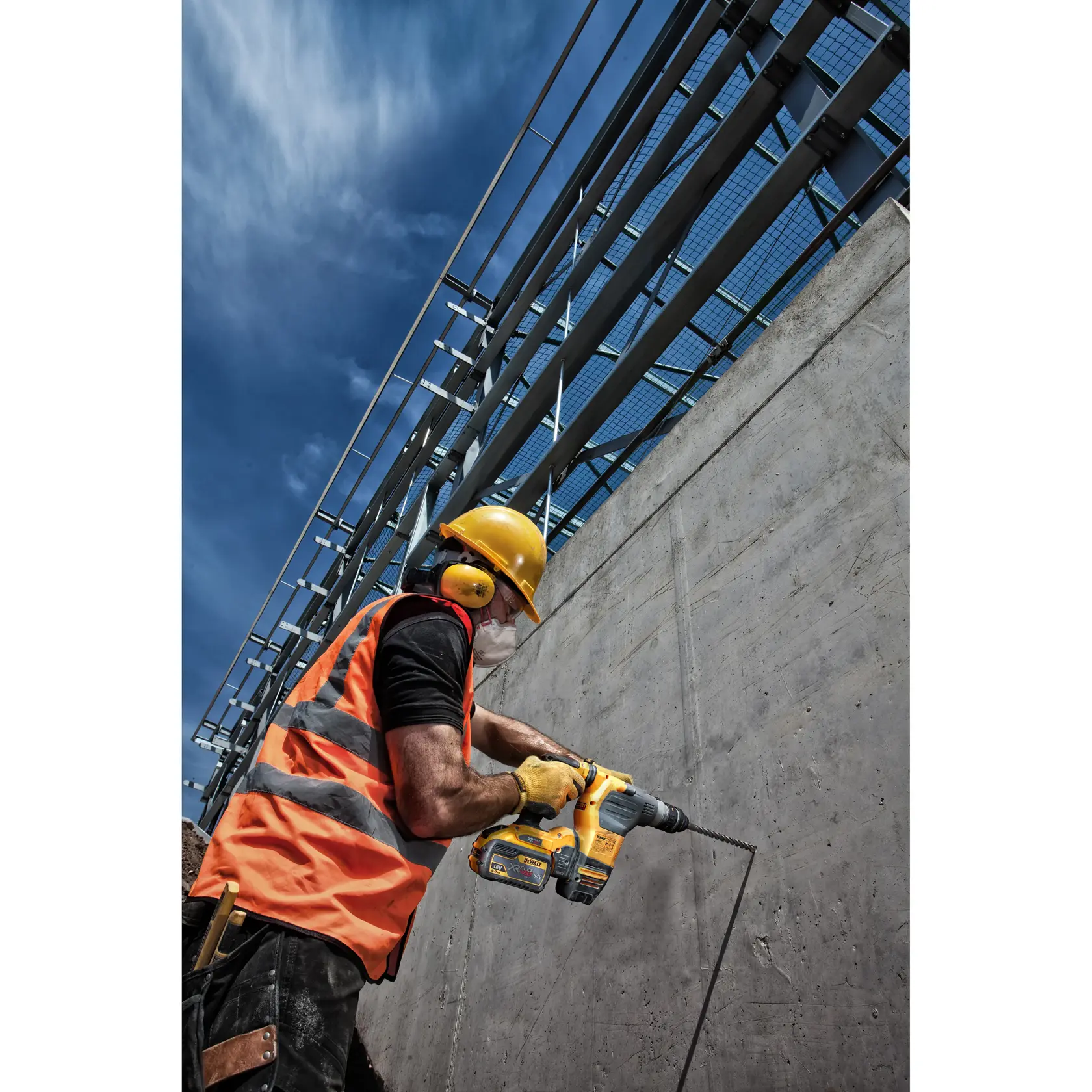 Worker with safety gear uses a DEWALT drill on a concrete wall at a construction site under a blue sky.