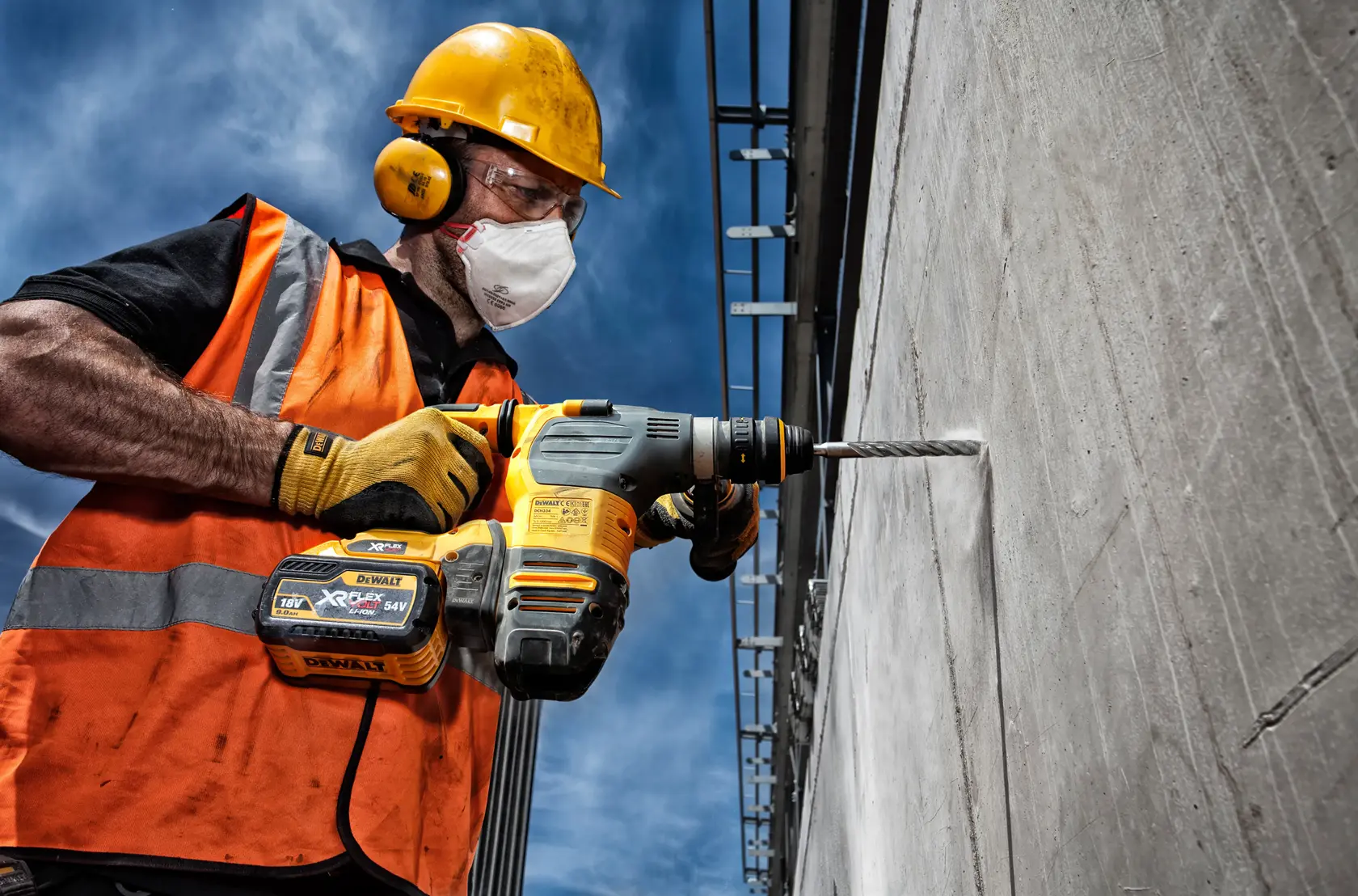 Worker uses DEWALT hammer drill to drill into a concrete wall outdoors, wearing safety gear and orange vest.
