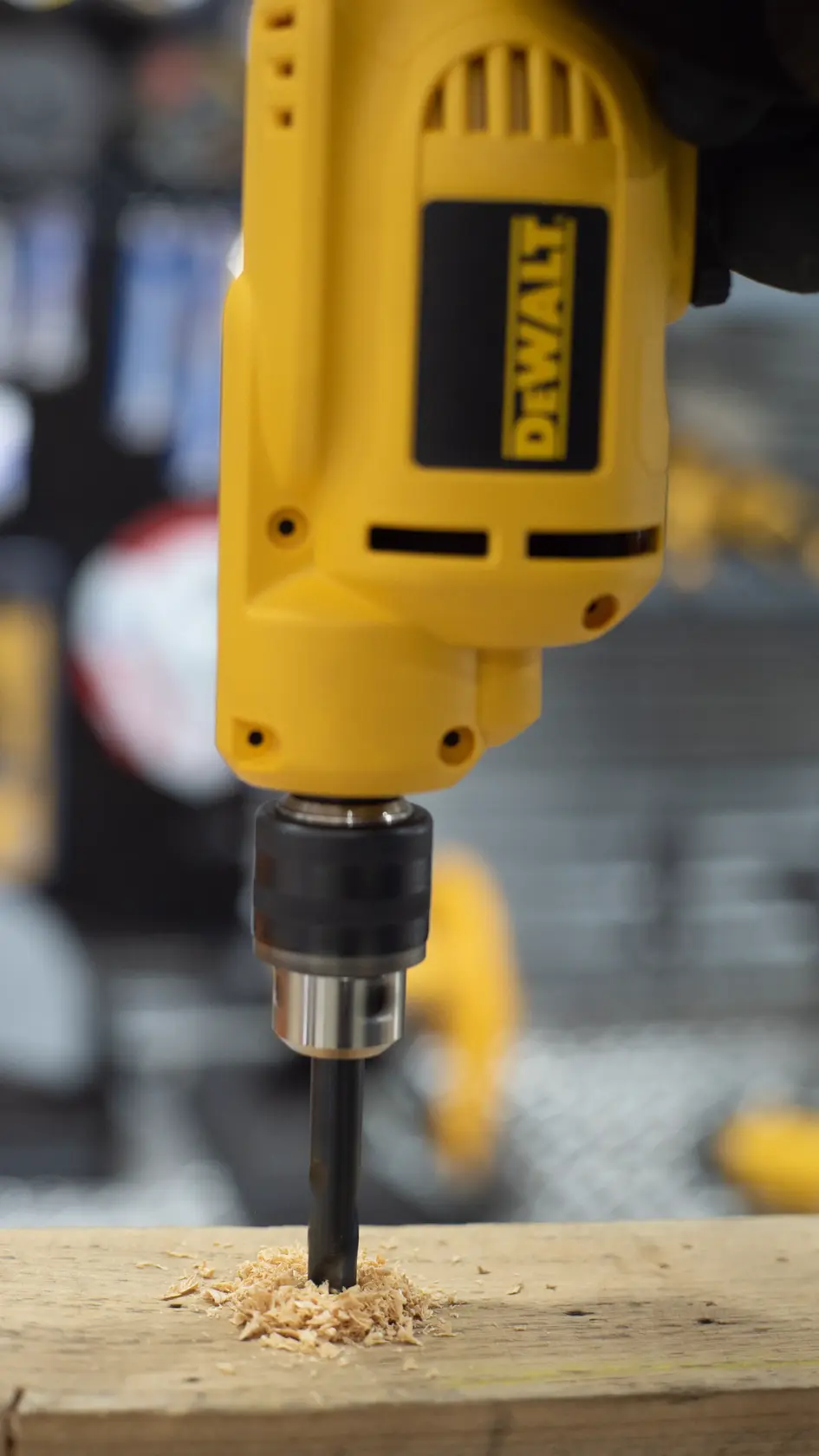 A close-up view of a yellow DEWALT electric drill being used to drill into a wooden plank, with wood shavings accumulating around the drill bit.
