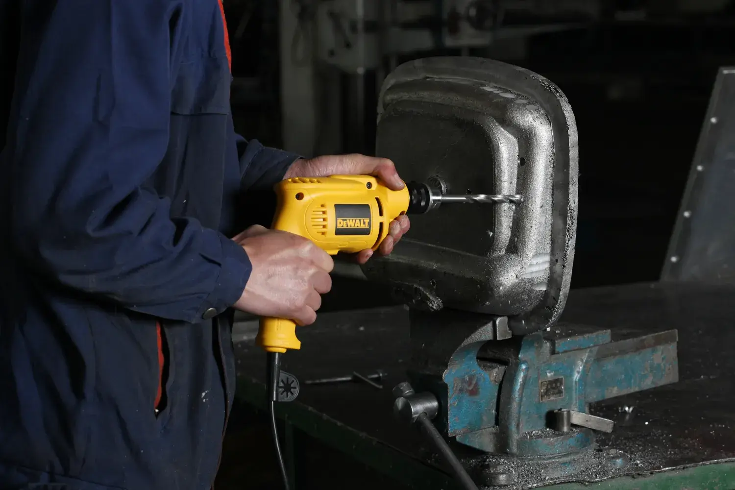 A person using a yellow DEWALT electric drill to work on a metal object secured in a vise on a workbench.