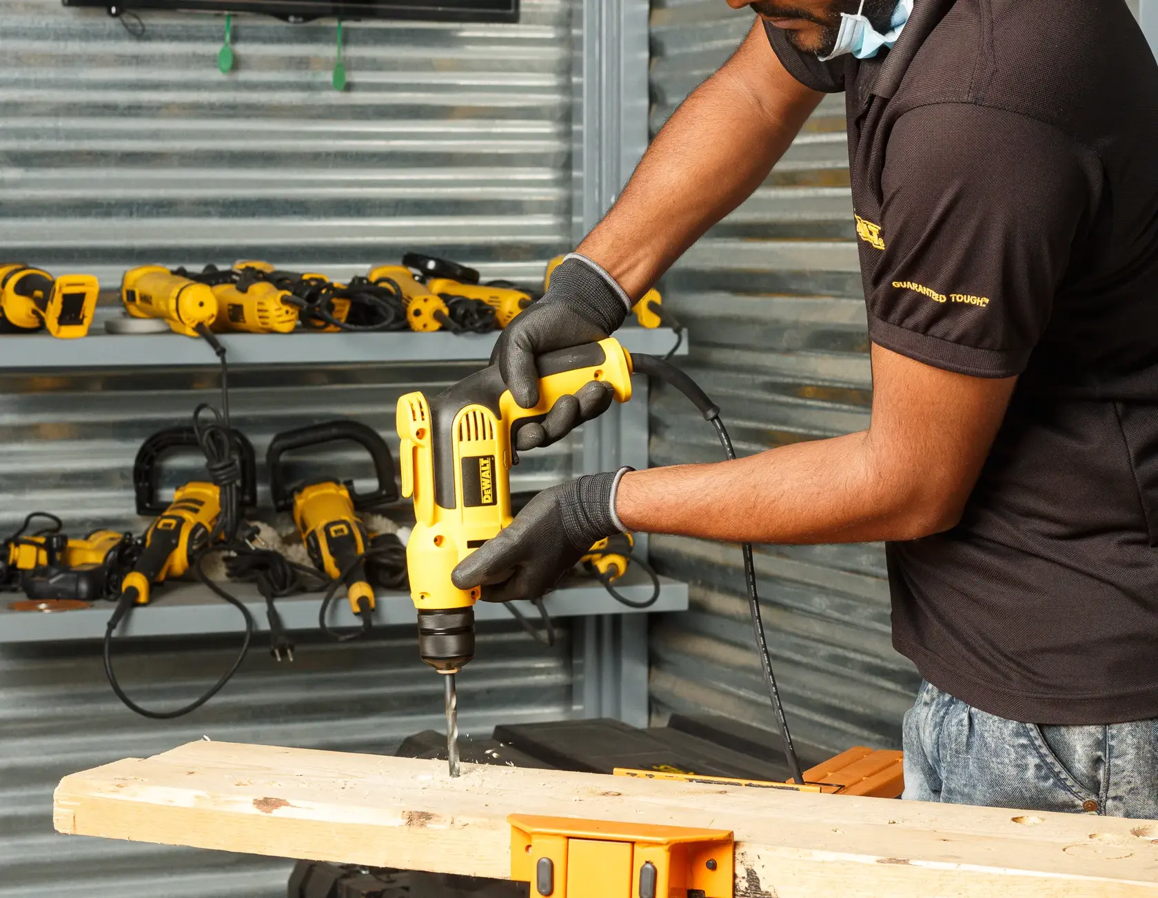 A person wearing gloves drills into a wooden plank using a yellow DEWALT electric drill, with other DEWALT power tools visible on shelves in the background.