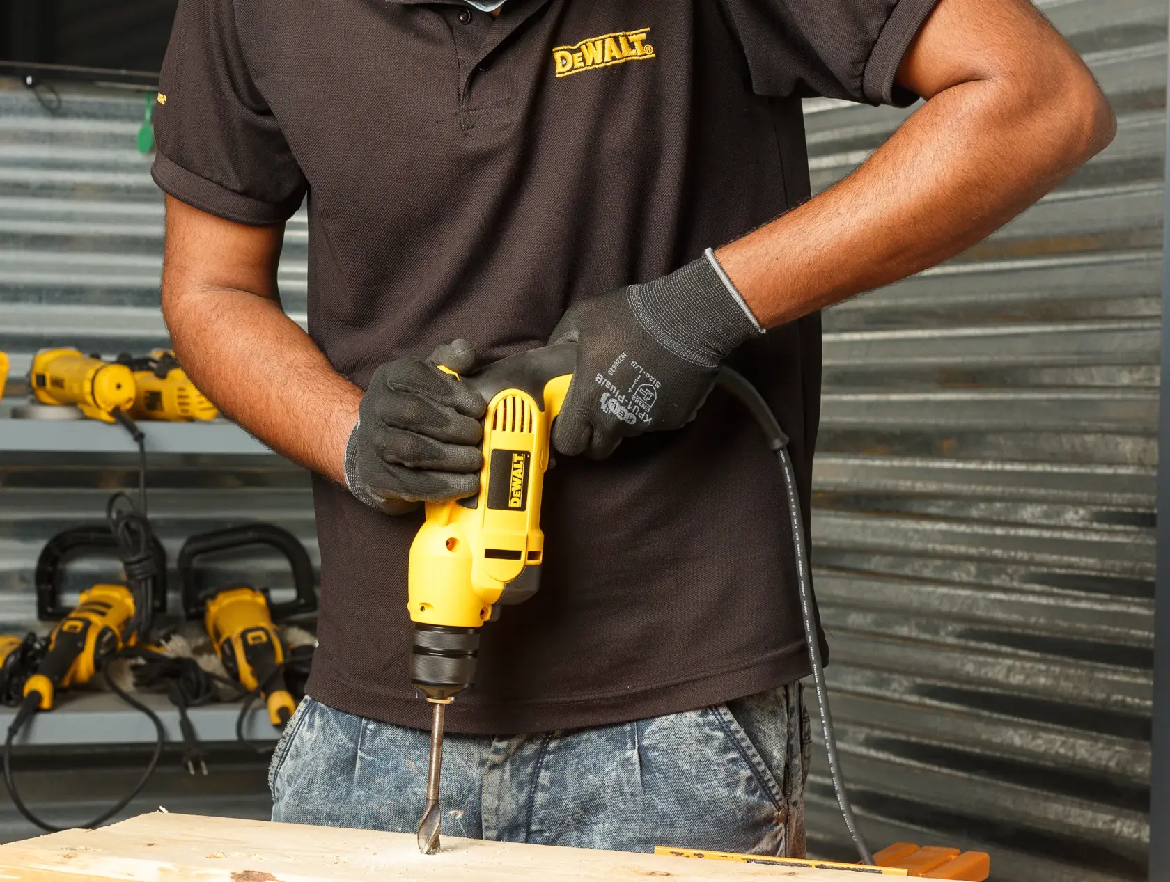 Person wearing gloves and a DEWALT shirt using a yellow DEWALT electric drill (model DWD112S) to drill into a wooden surface. Several DEWALT tools are visible in the background on shelves.