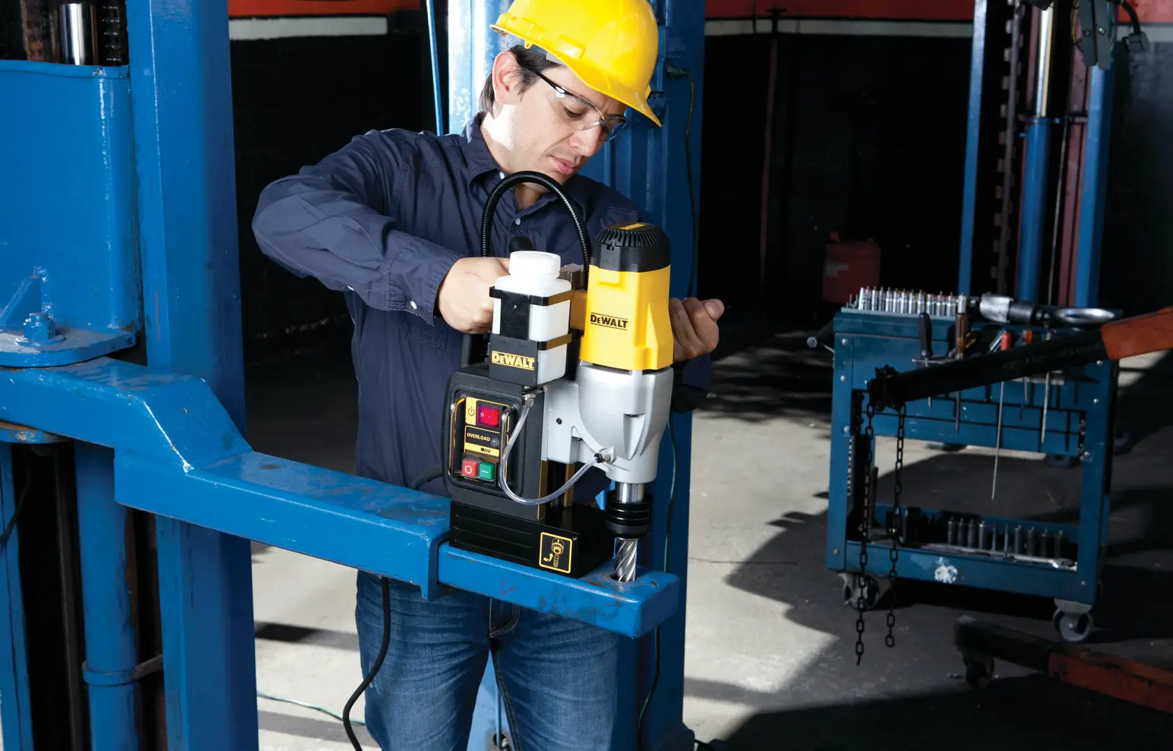 A person wearing a yellow hard hat operates a DEWALT DWE1622K magnetic drill press on a blue metal beam in an industrial workshop setting. The individual's face is blurred for privacy.