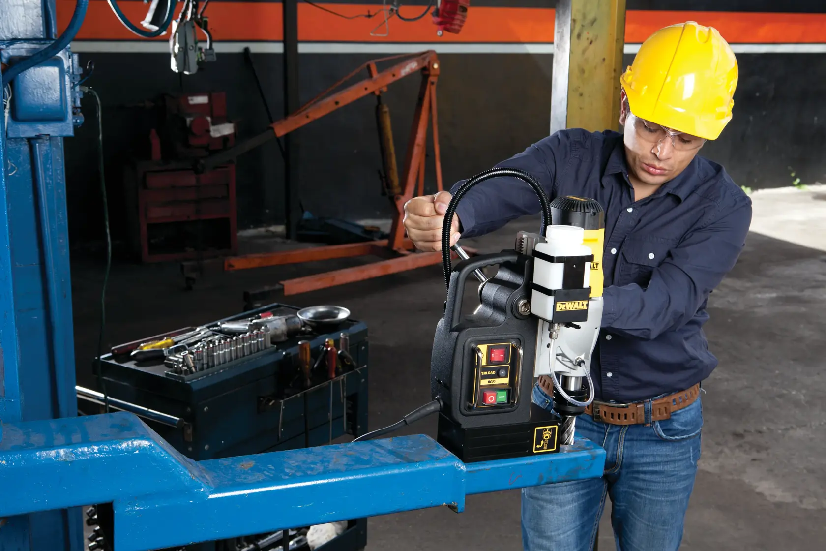 A person wearing a yellow safety helmet operates a DEWALT DWE1622K magnetic drill on a metal beam in an industrial workshop. Nearby, there is a toolbox with various tools. The person's face is blurred for privacy.